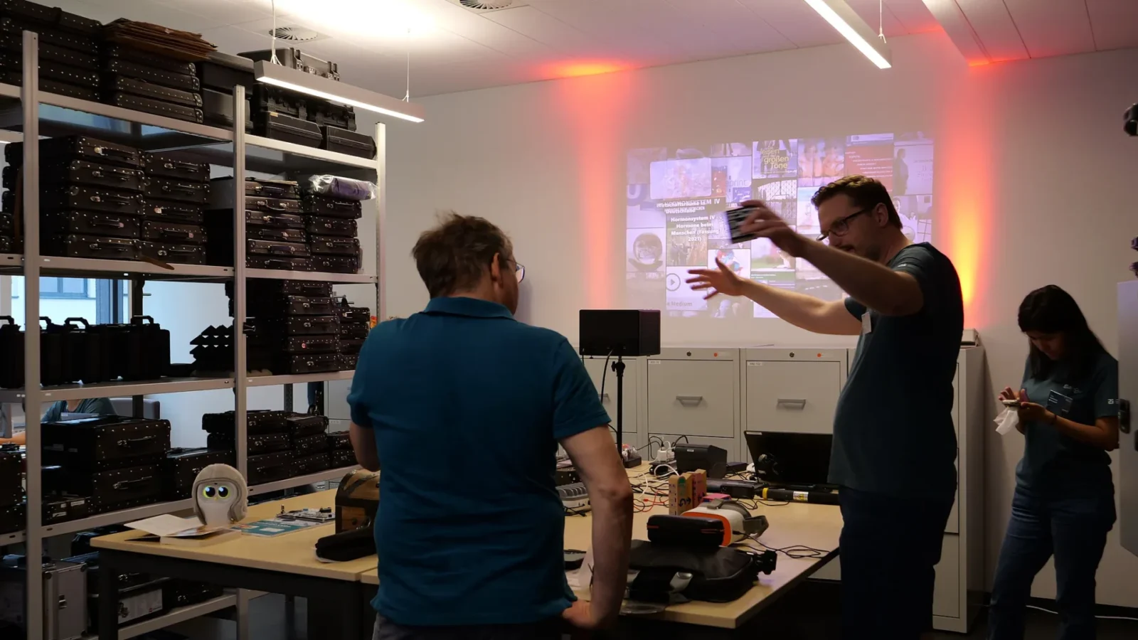 Mitarbeiter erklärt vor einer Beamer-Projektion mit Medienangeboten, dahinter Regale voller Transportkoffer