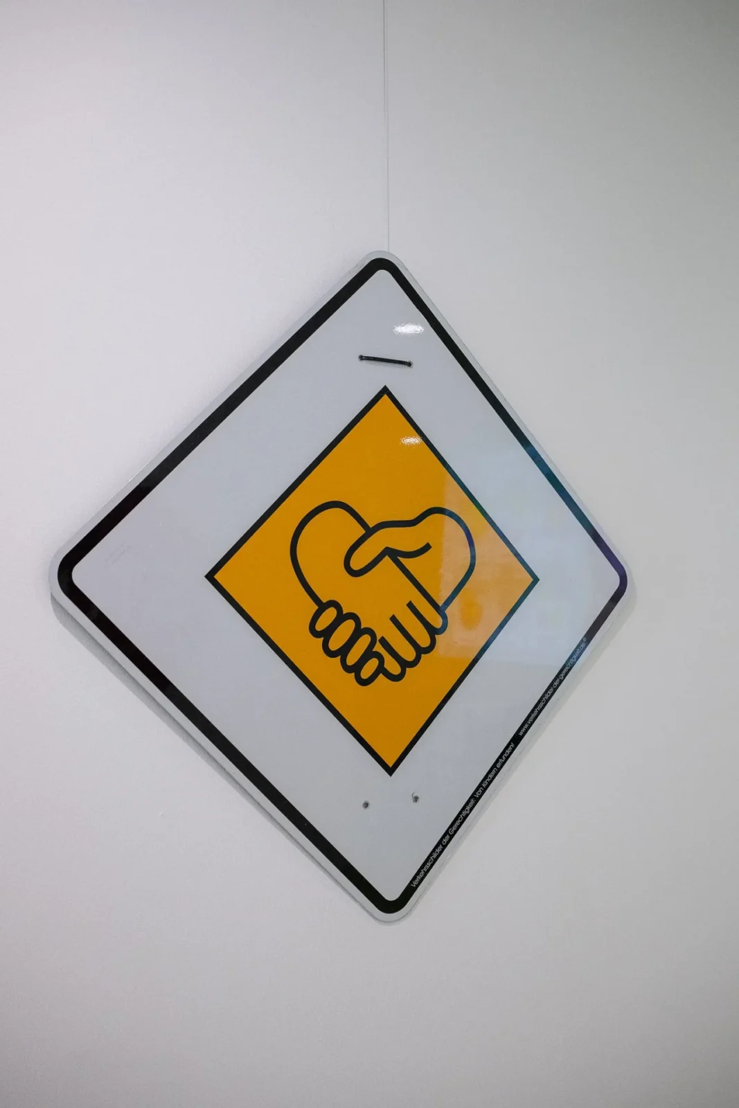 Kunst-Verkehrsschild als gelbe Raute mit Handschlag-Symbol in Herzform an weißer Wand