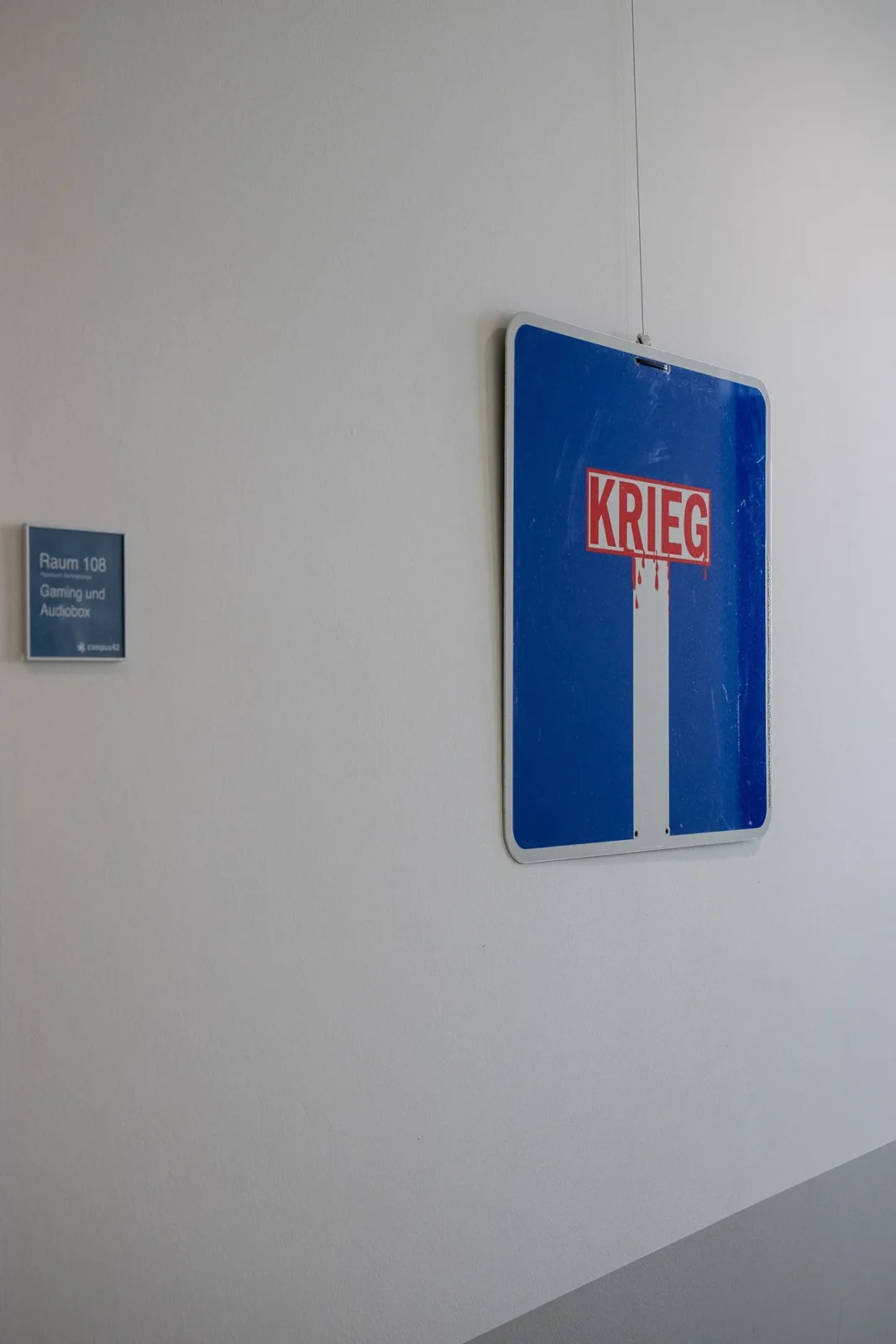 Blaues Kunst-Verkehrsschild mit rotem Schriftzug „KRIEG" neben Türschild „Raum 108 – Gaming und Audiobox"