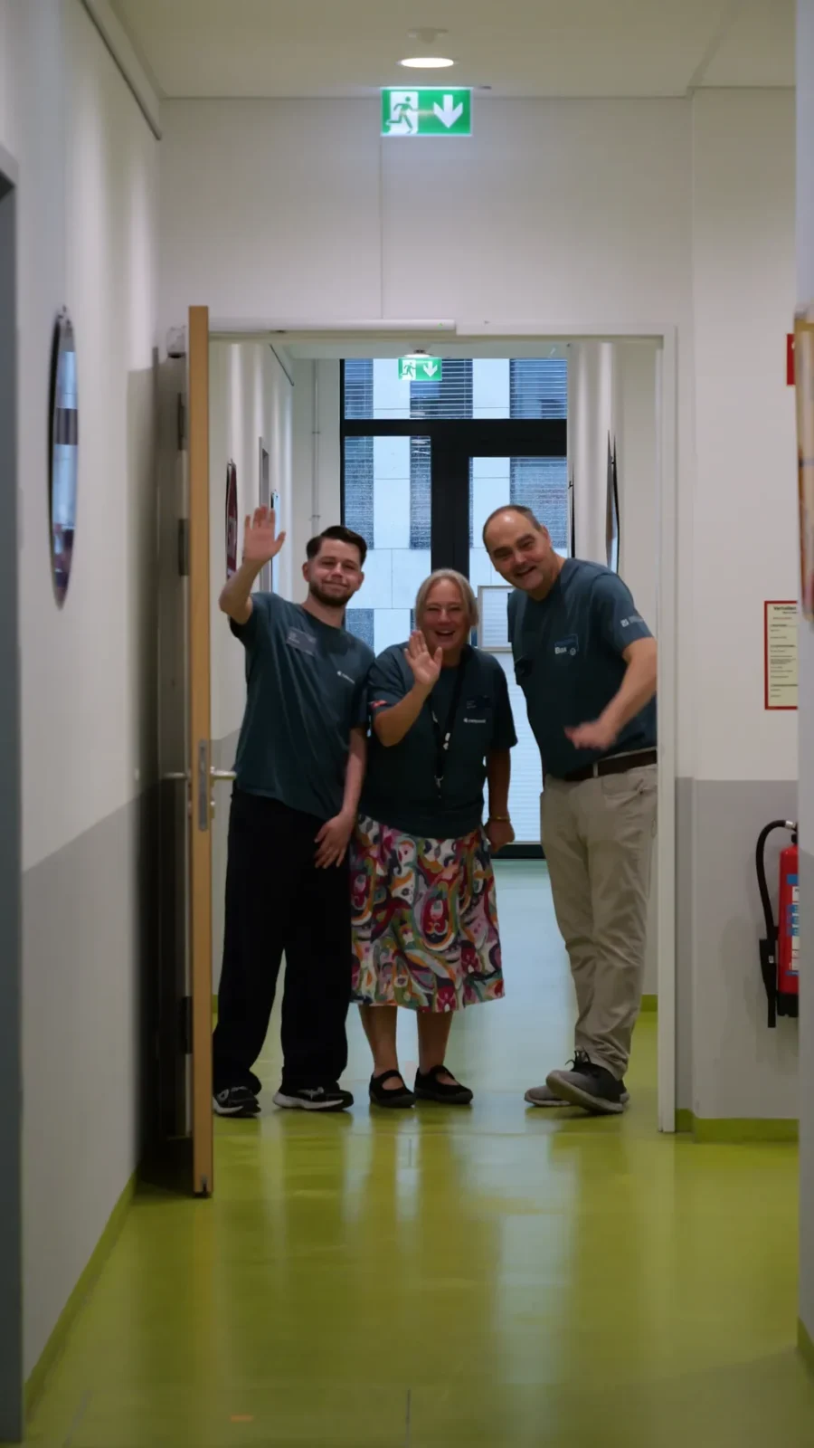 Drei Teammitglieder in campus42-Shirts winken lächelnd im Türrahmen eines Flurs