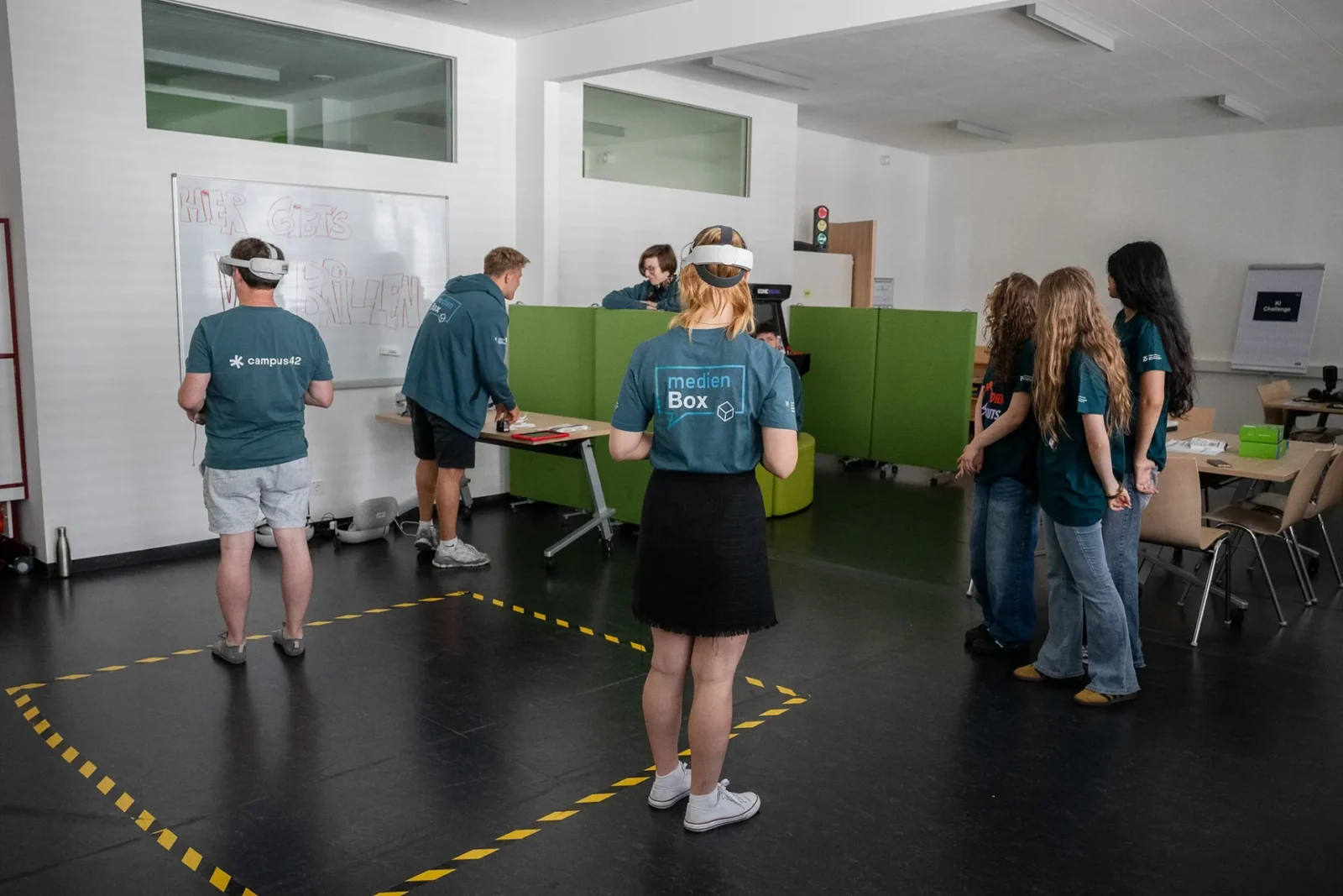 Person mit VR-Brille im gelb markierten Spielfeld, weitere Gäste in medienBox-Shirts beobachten die VR-Station