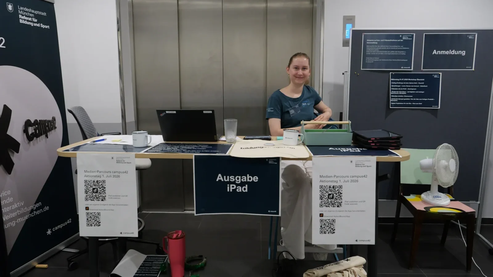 Mitarbeiterin am Anmeldetisch mit Schildern „Ausgabe iPad" und QR-Codes zum Medien-Parcours