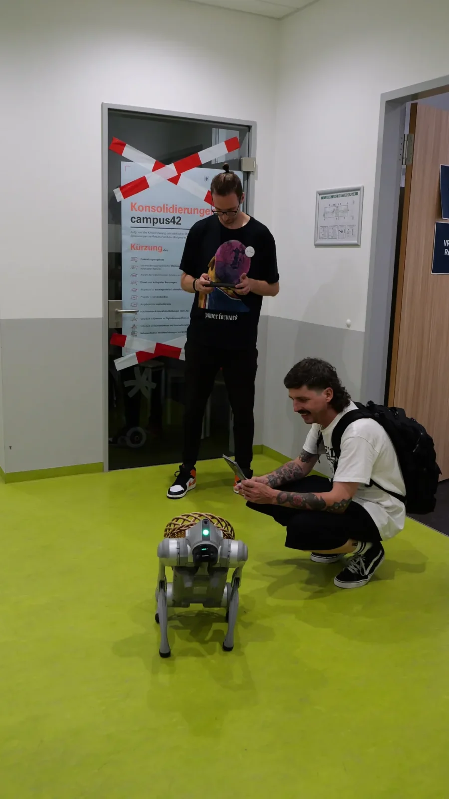 Zwei Besucher steuern den Roboterhund per Tablet vor einer mit Absperrband gekreuzten Tür
