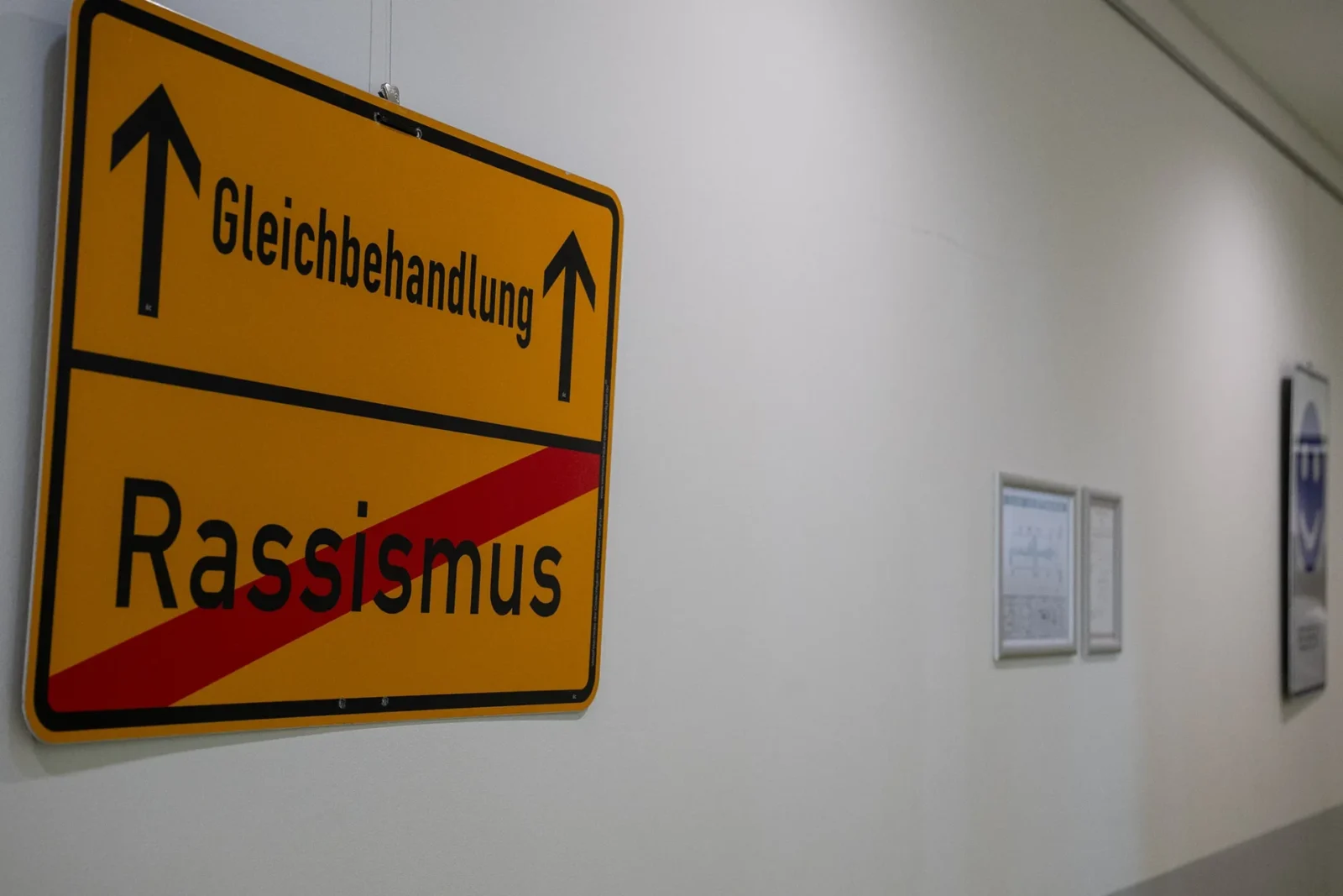 Gelbes Ortsschild im Flur: „Gleichbehandlung" mit Pfeilen, darunter durchgestrichenes „Rassismus"