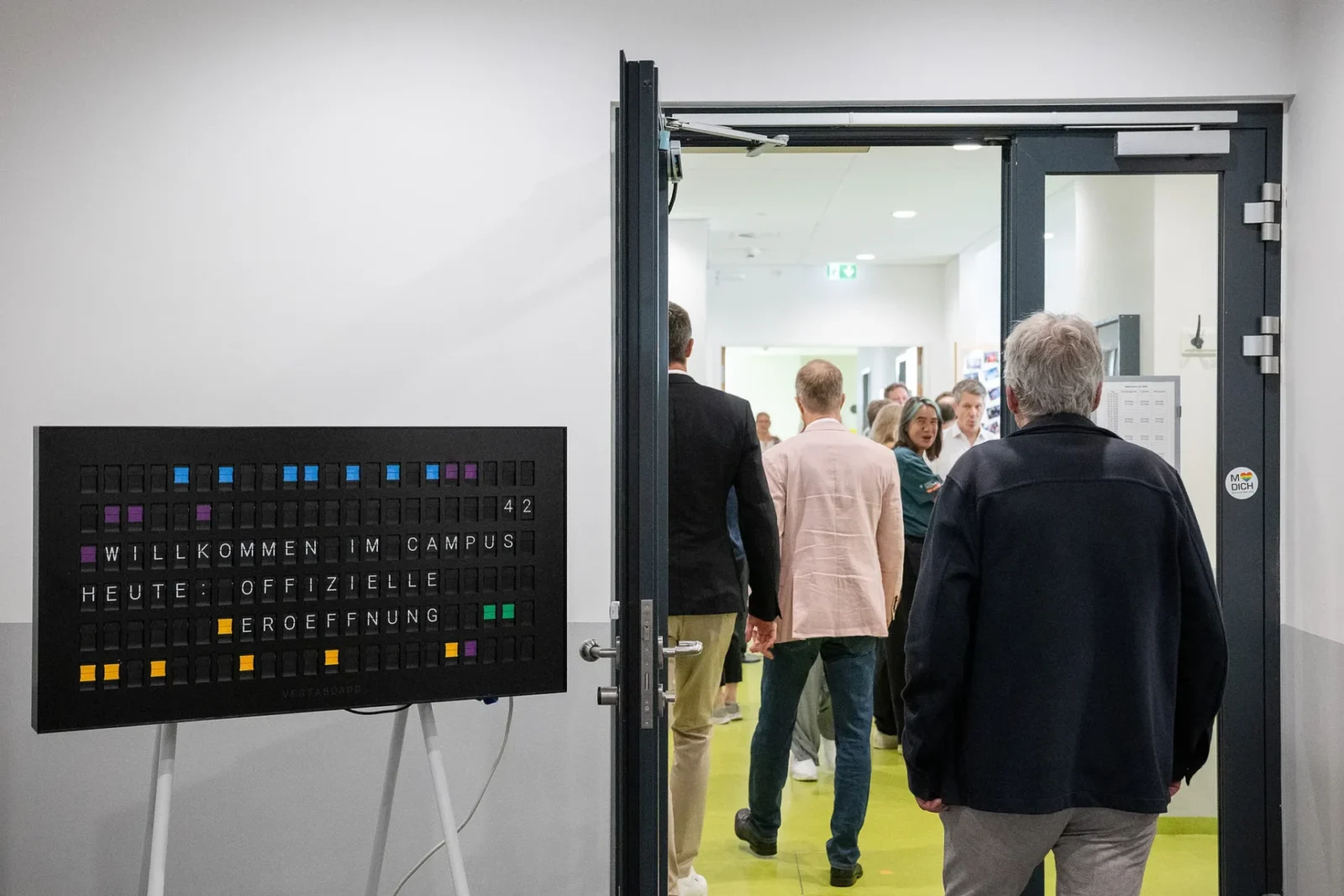 Anzeigetafel „Willkommen im Campus 42 – Heute: Offizielle Eröffnung" neben offener Tür, durch die Gäste strömen