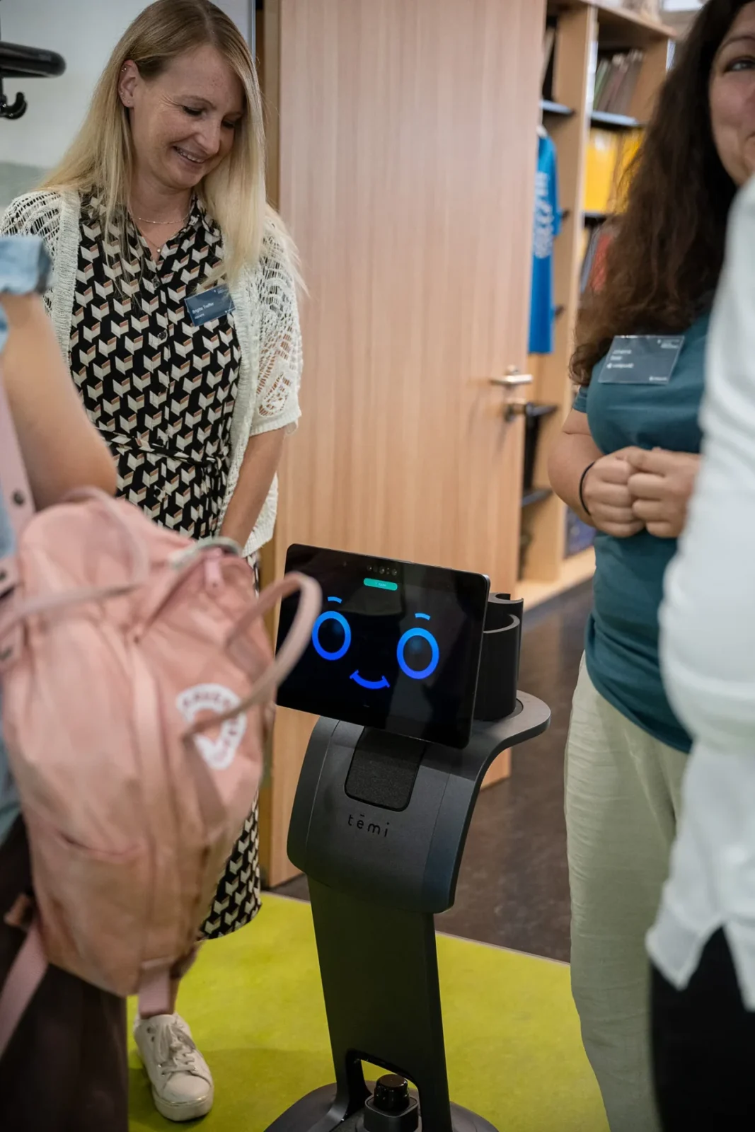 Zwei Besucherinnen betrachten schmunzelnd den Serviceroboter temi mit blauem Smiley-Gesicht