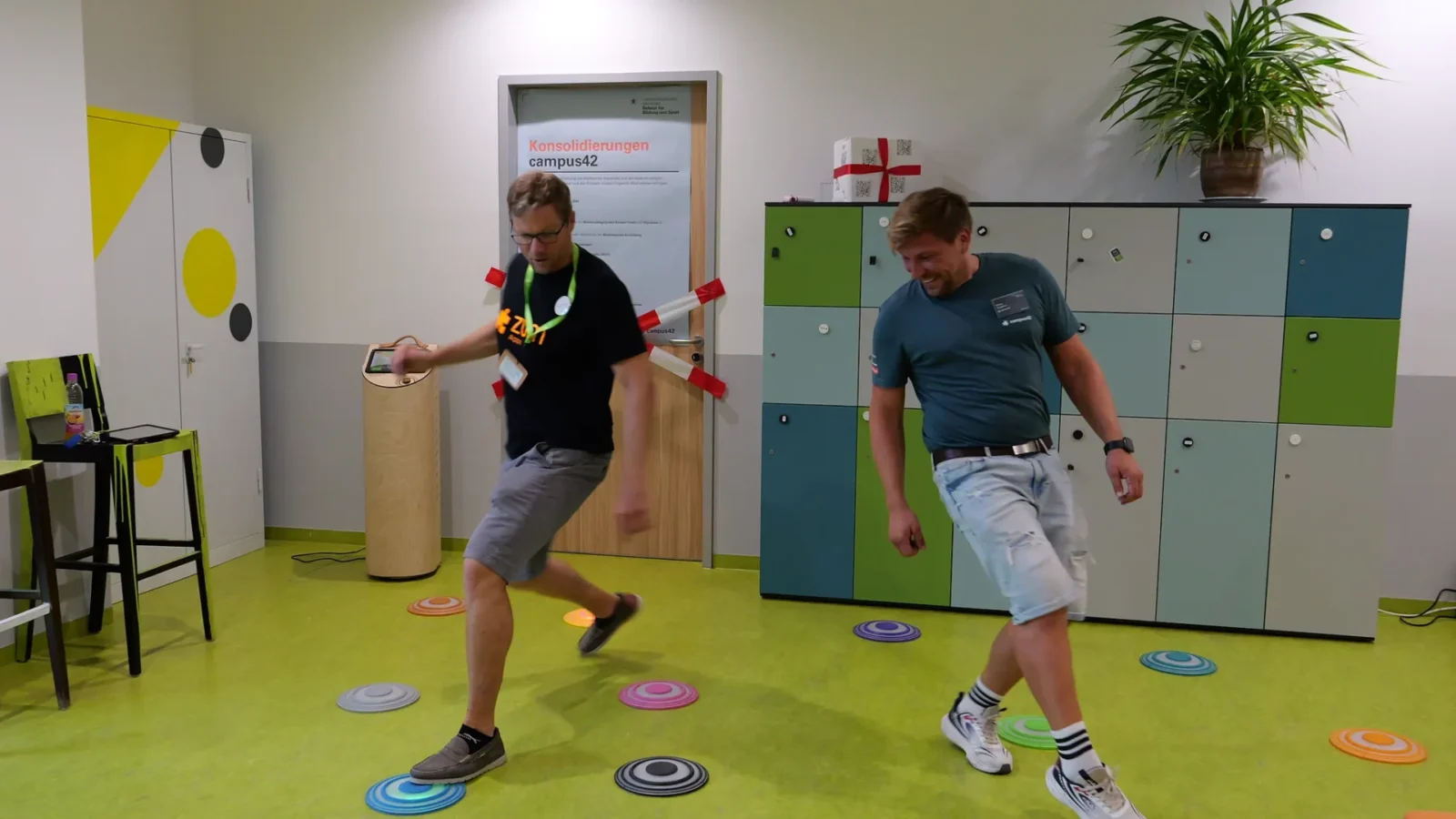 Zwei Männer balancieren auf bunten Bodenmarkierungen bei einem Bewegungsspiel im Flur