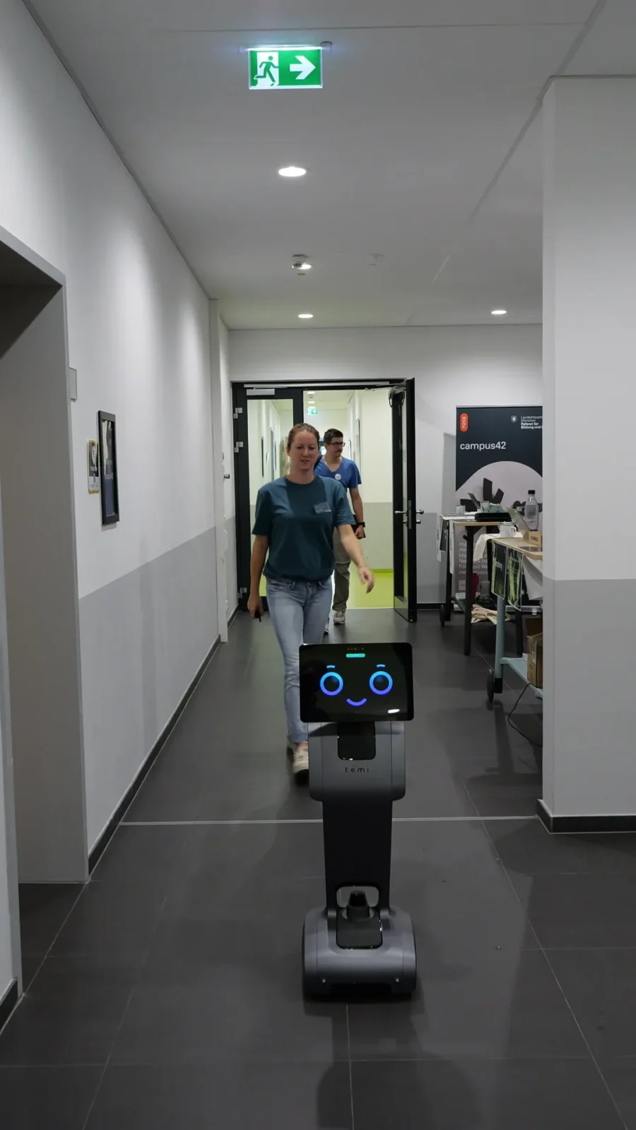 Serviceroboter temi mit lächelndem Display fährt durch den Flur, rechts campus42-Rollup