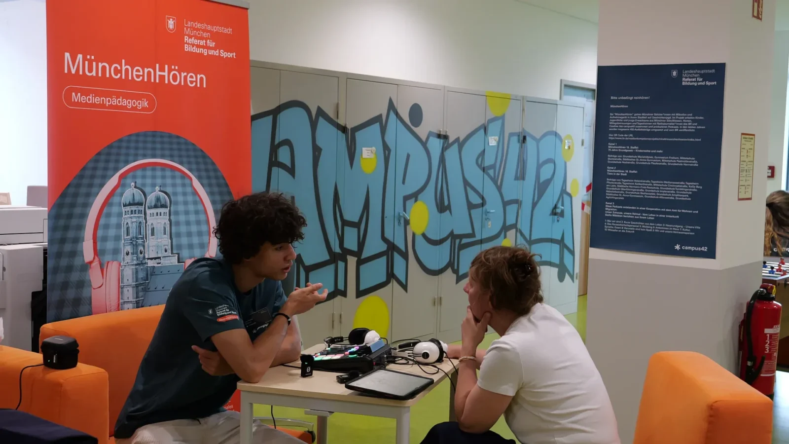 Podcast-Gespräch in Sesseln vor der campus42-Graffitiwand, Rollup „MünchenHören" links