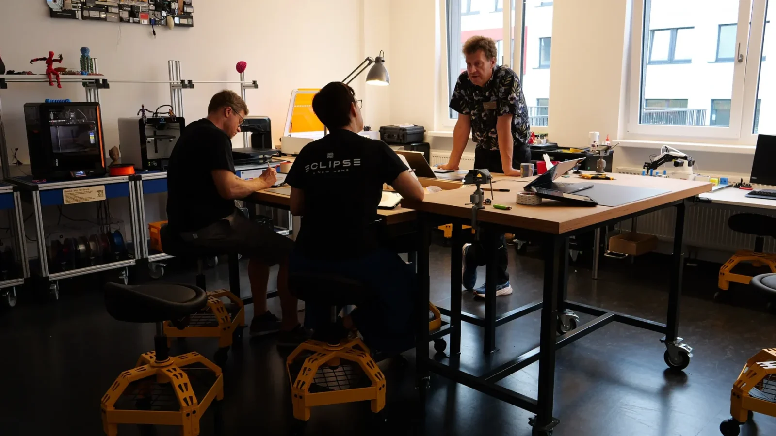 Makerspace mit 3D-Druckern und Lasercutter, drei Personen an der Werkbank mit Laptops