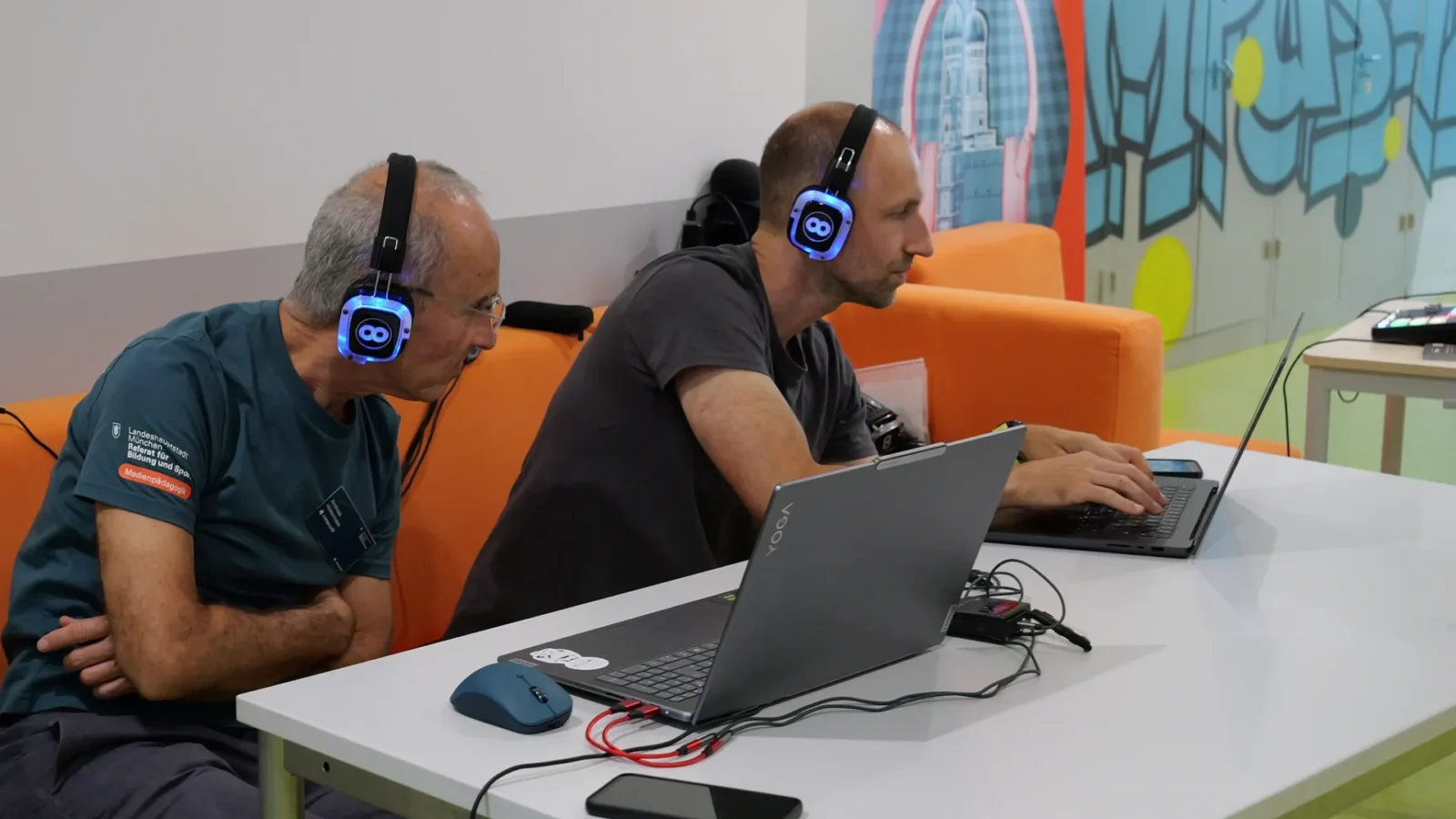 Zwei Männer mit blau leuchtenden Kopfhörern arbeiten am Laptop vor dem MünchenHören-Motiv