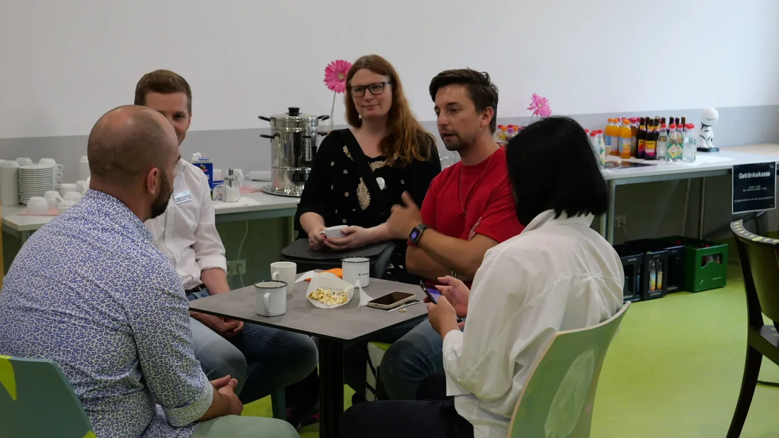 Fünf Personen mit Tassen und Popcorn am Tisch im Cafébereich, dahinter Schild „Getränkekasse"
