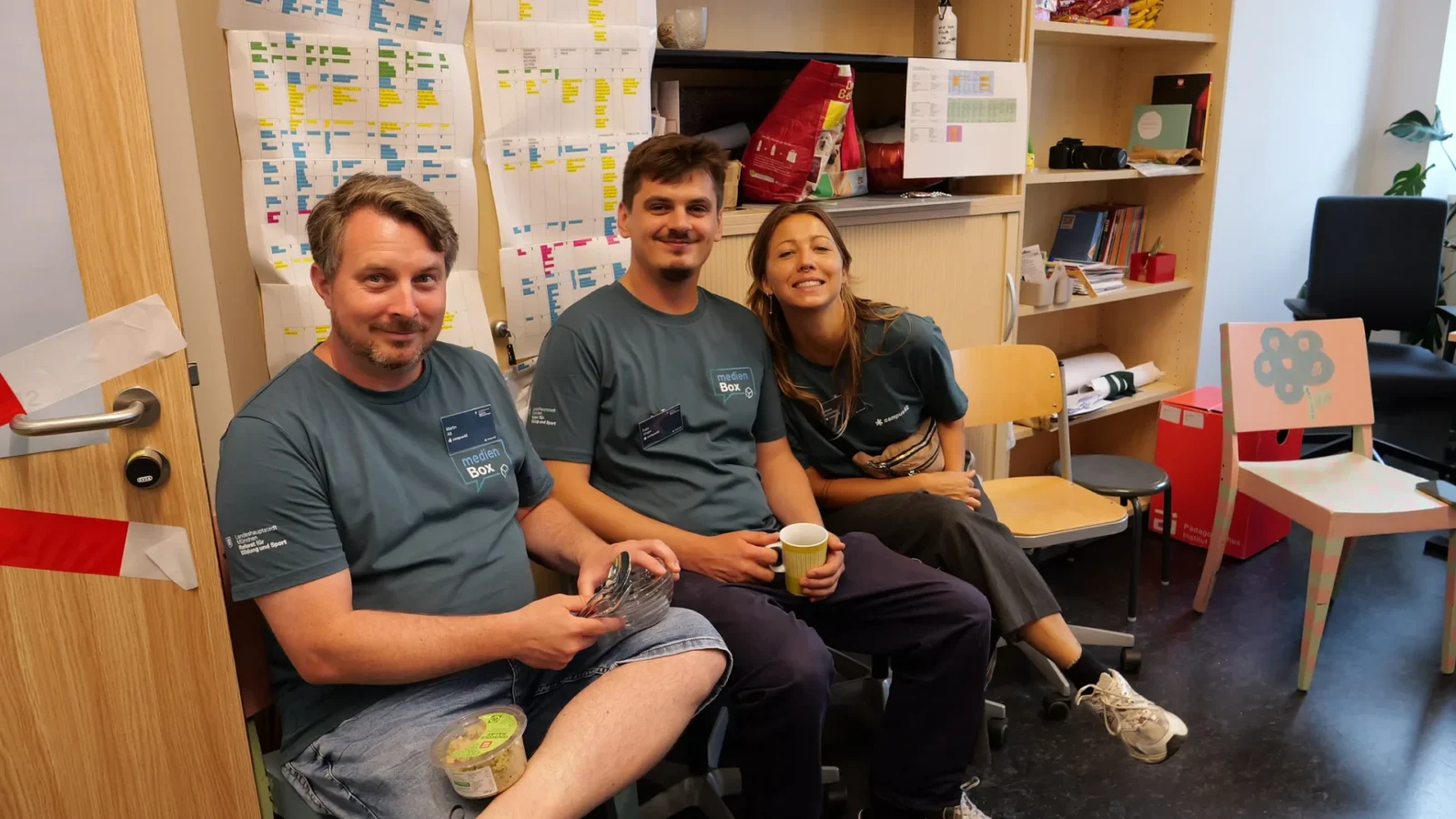 Drei Teammitglieder in medienBox-Shirts sitzen lachend mit Kaffeebecher vor einer Planungswand