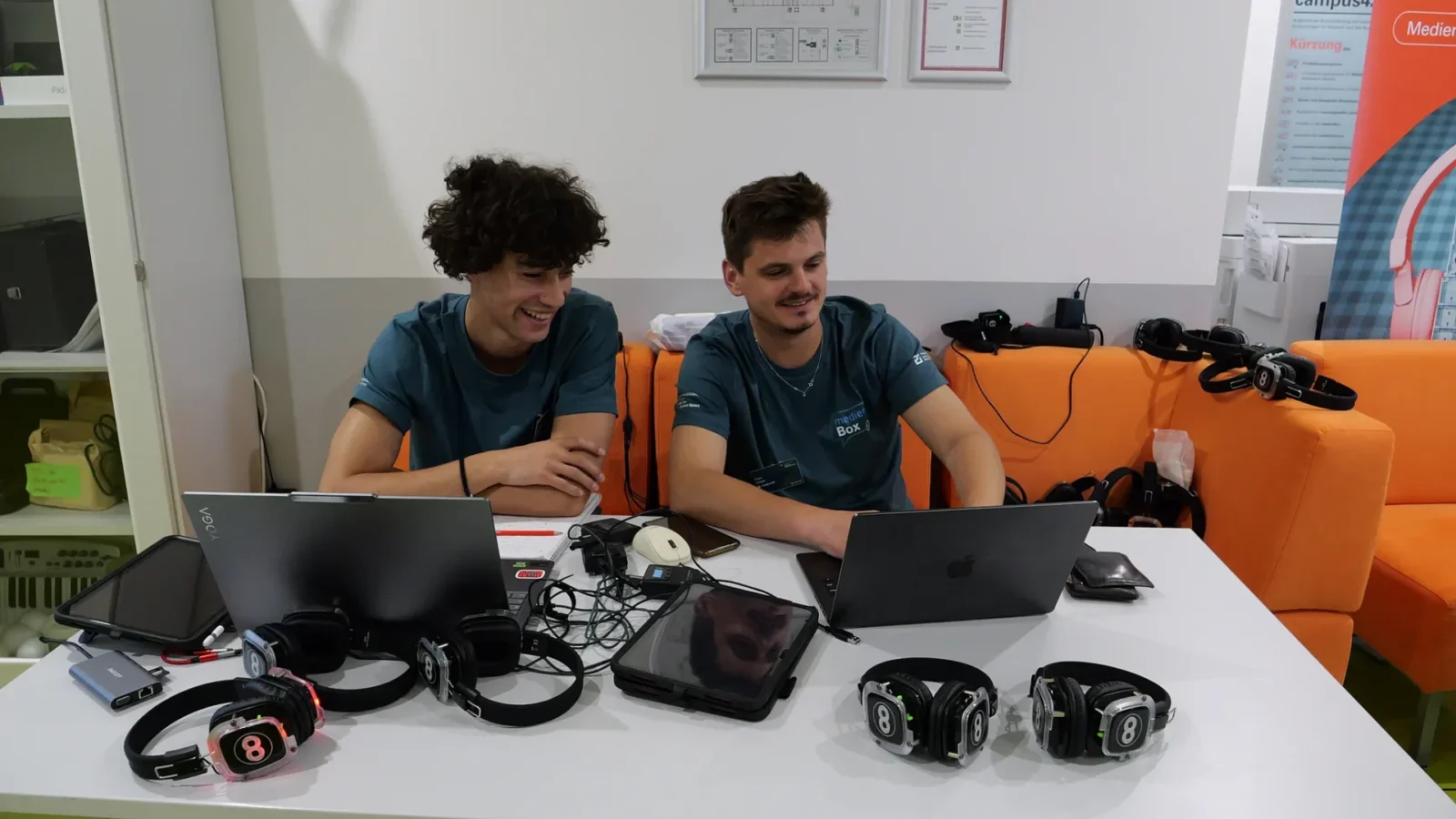 Zwei Mitarbeitende in medienBox-Shirts arbeiten an Laptops, davor mehrere Funkkopfhörer