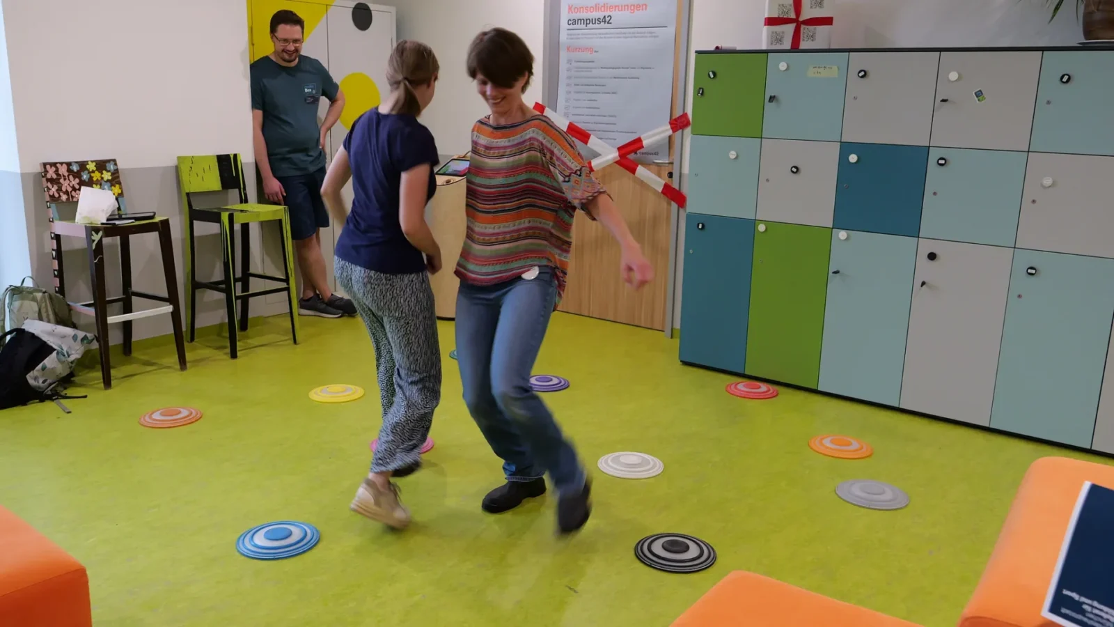 Zwei Frauen tanzen auf einem Bodenparcours aus bunten Balance-Punkten, ein Mitarbeiter schaut zu