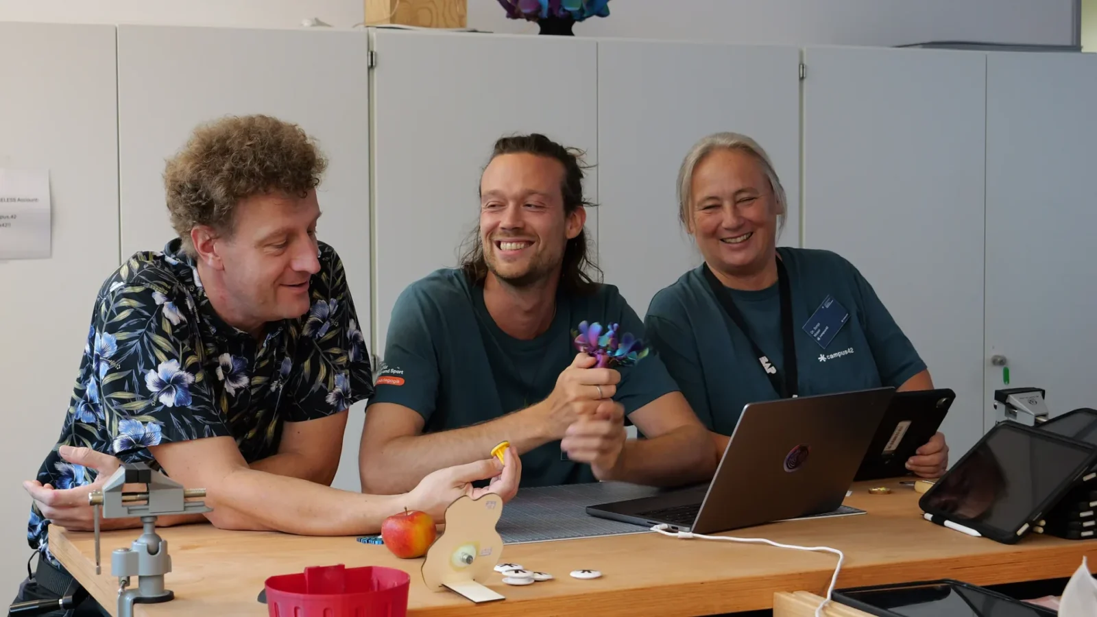 Drei Personen lachen an einem Makerspace-Tisch mit Laptop, Schraubstock und Holzteilen