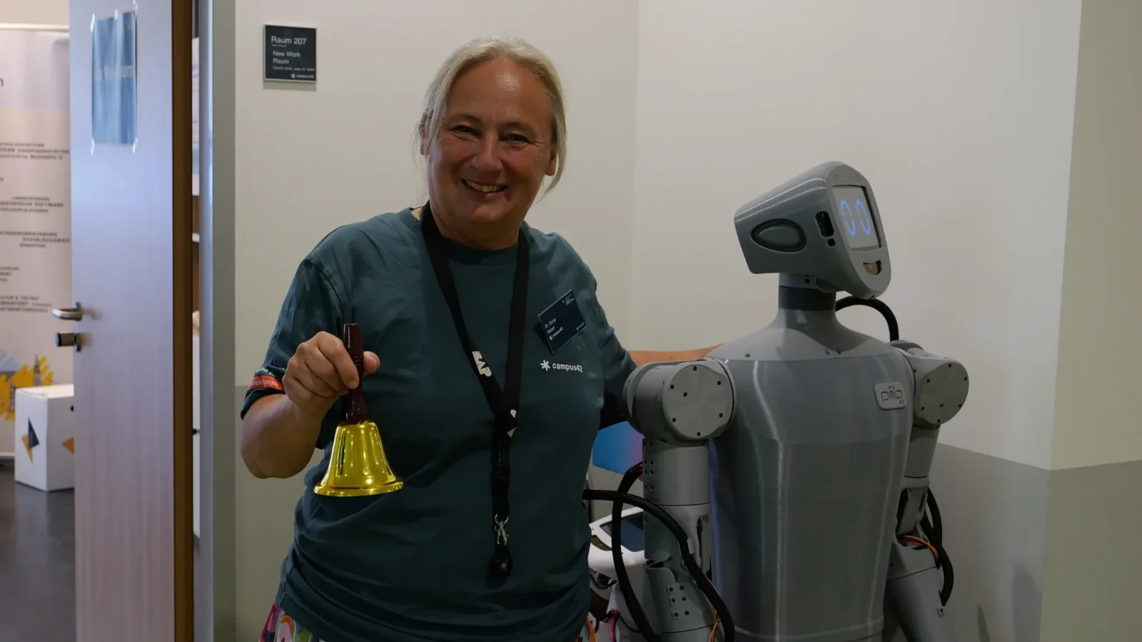 Mitarbeiterin läutet eine Handglocke neben einem 3D-gedruckten Roboter vor Raum 207 „New Work Raum"