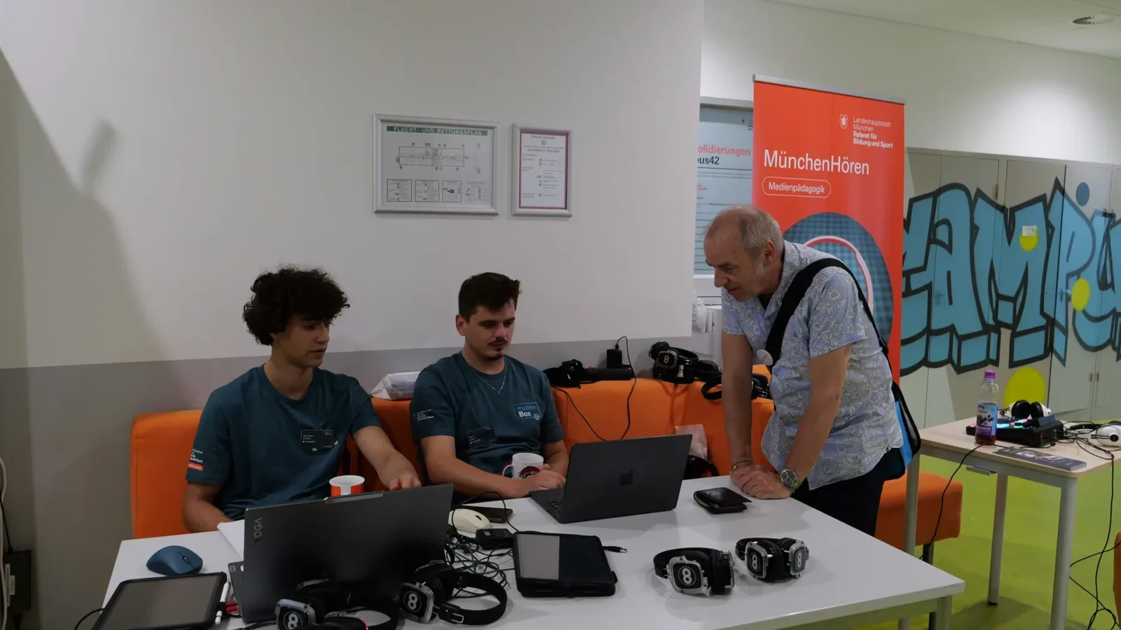 Zwei junge Mitarbeiter am Laptop mit Gaming-Headsets, daneben Rollup „MünchenHören"