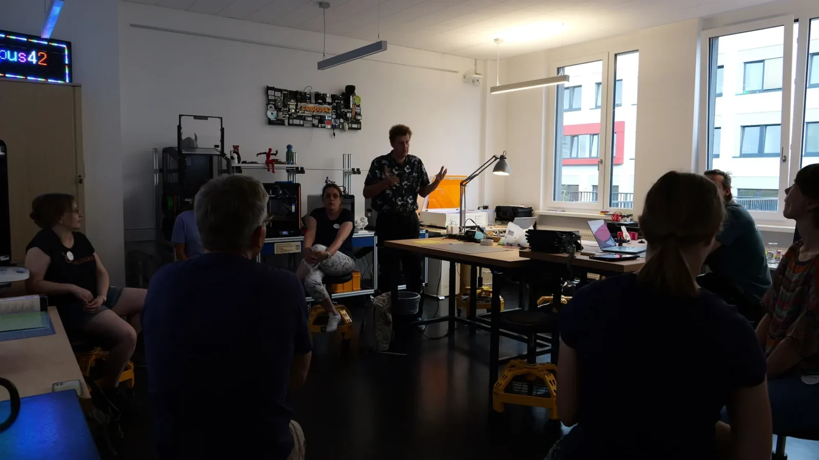 Vortrag im Makerspace vor 3D-Druckern und LED-Laufschrift „campus42", Zuhörende auf Rollhockern