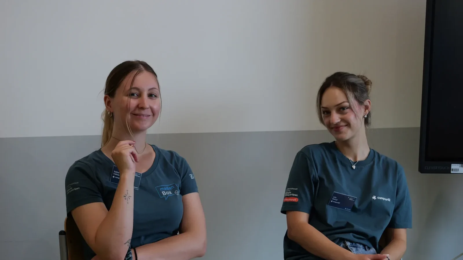 Zwei Mitarbeiterinnen in campus42- und medienBox-Shirts sitzen lächelnd vor einem Touchdisplay