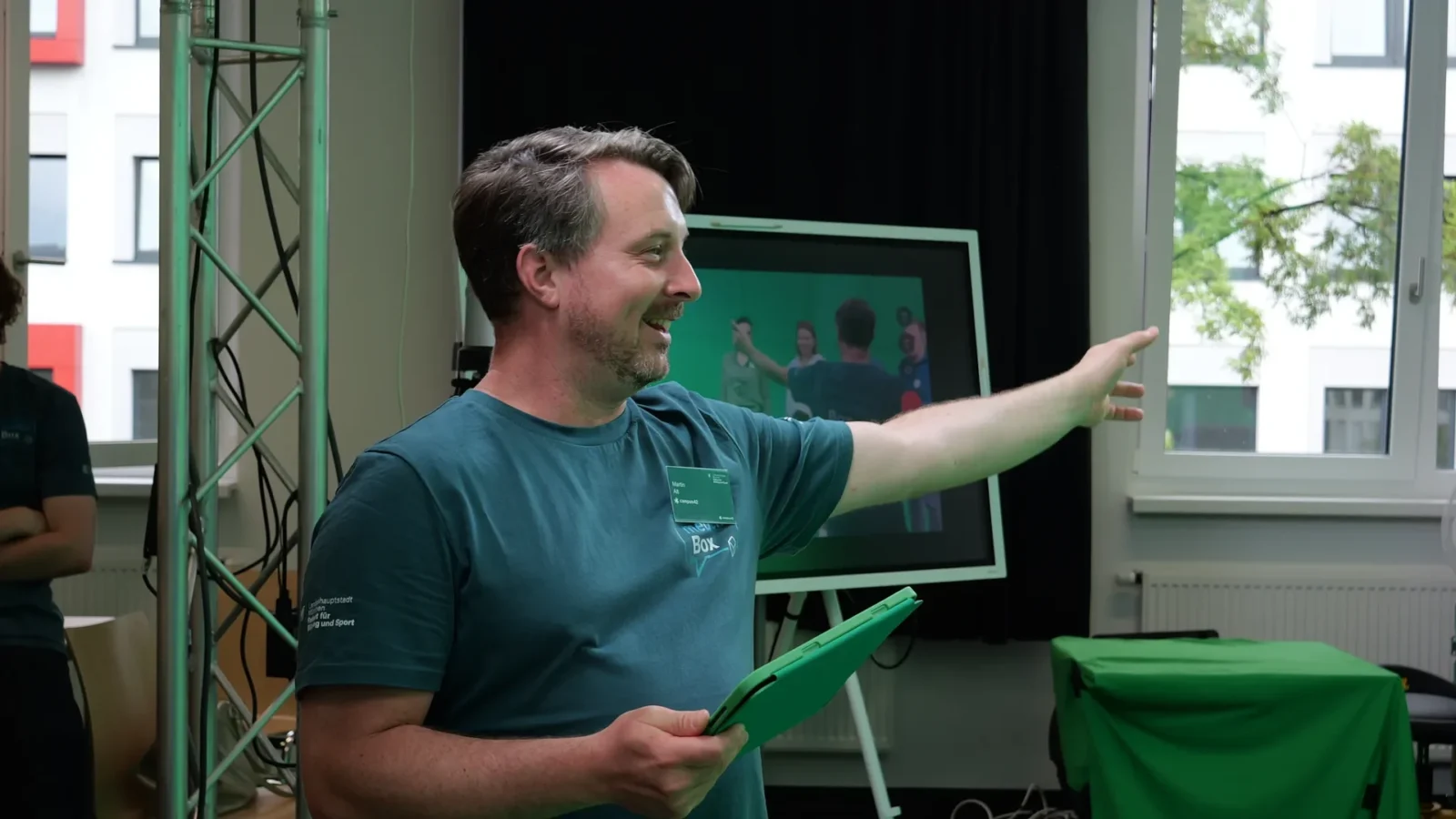 Mitarbeiter im medienBox-Shirt weist in Richtung Greenscreen-Studio, Tablet in der Hand
