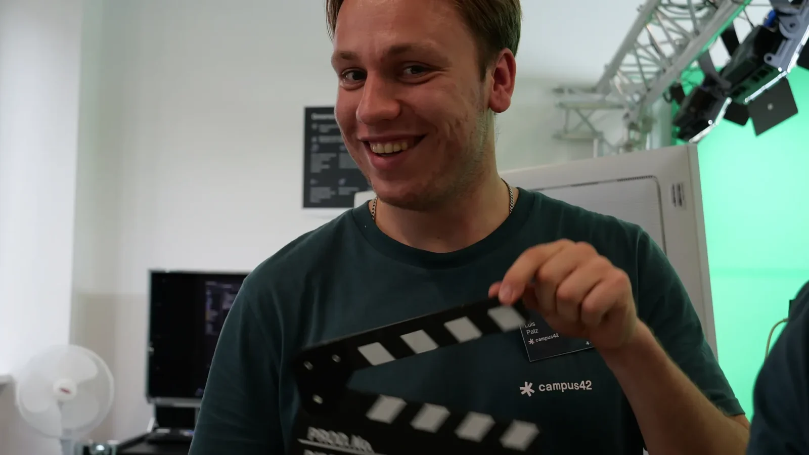Lachender Mitarbeiter im campus42-Shirt hält eine Filmklappe vor dem Greenscreen-Studio