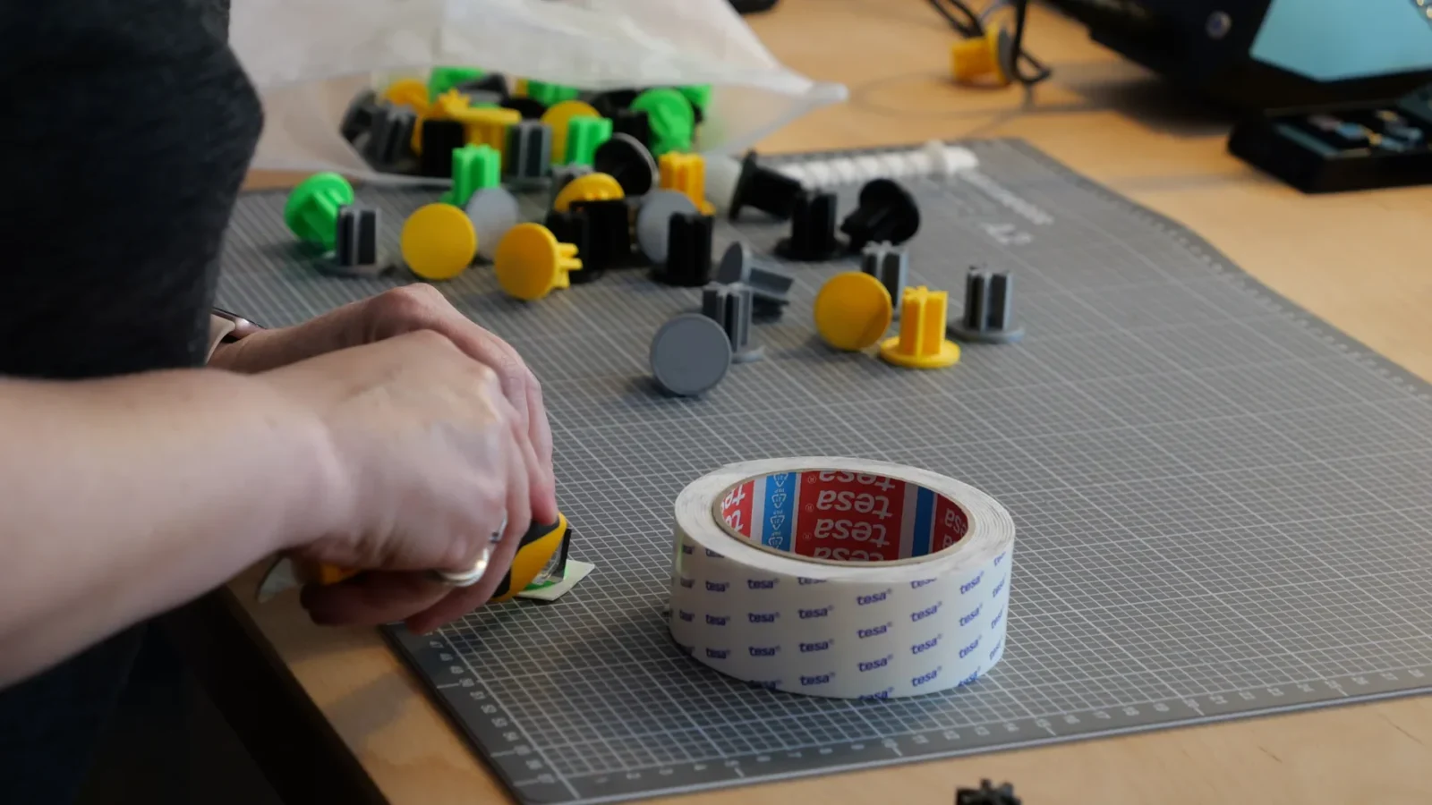 Hand schneidet mit Cuttermesser auf einer Schneidematte, daneben bunte 3D-gedruckte Bauteile