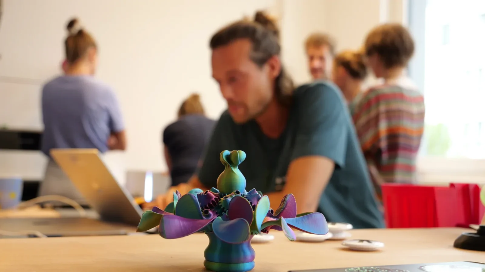 Schillerndes 3D-gedrucktes Blumenobjekt im Vordergrund, dahinter unscharf Menschen am Werktisch