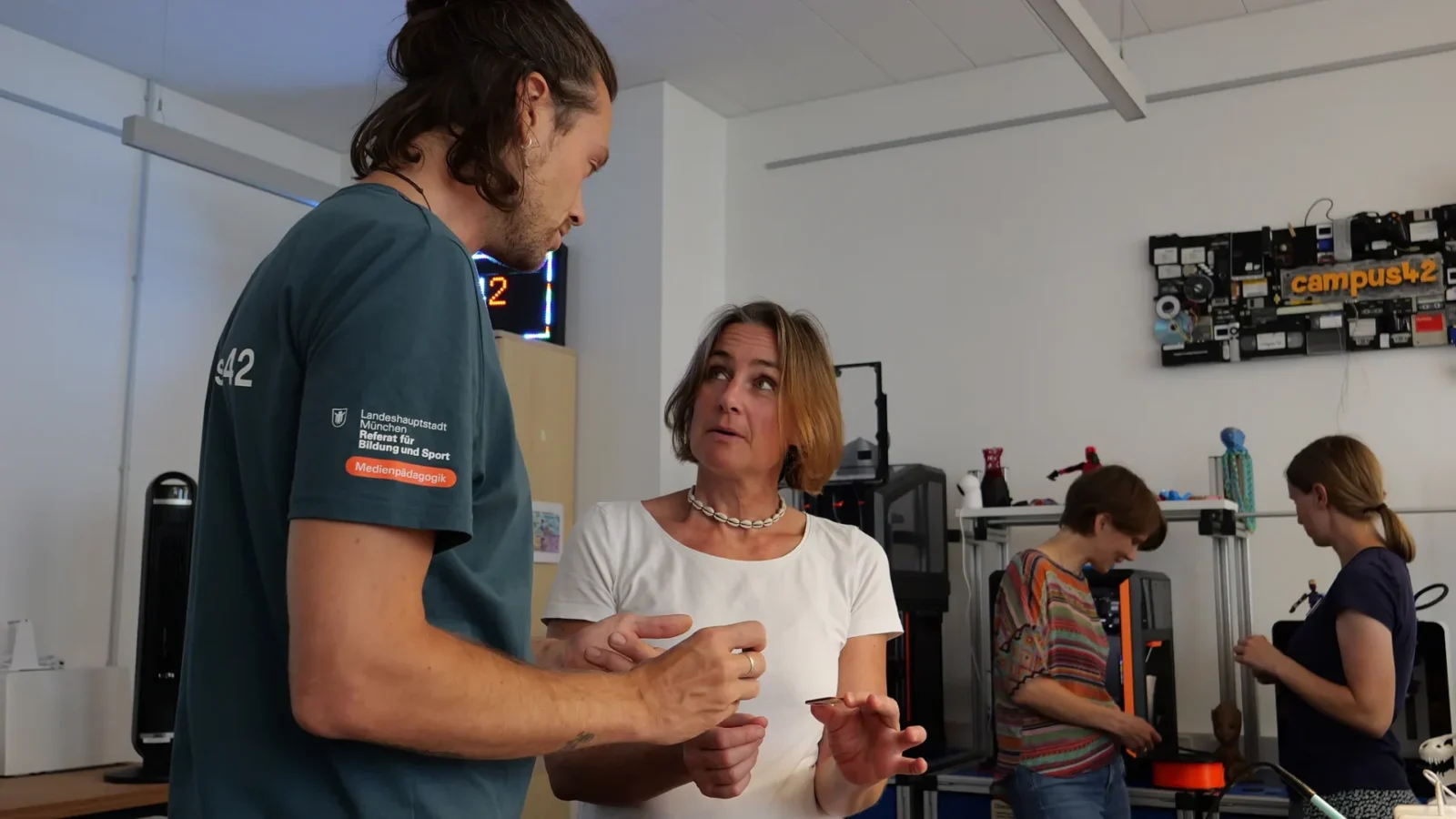 Gespräch im Makerspace vor 3D-Druckern, LED-Schriftzug campus42 an der Wand