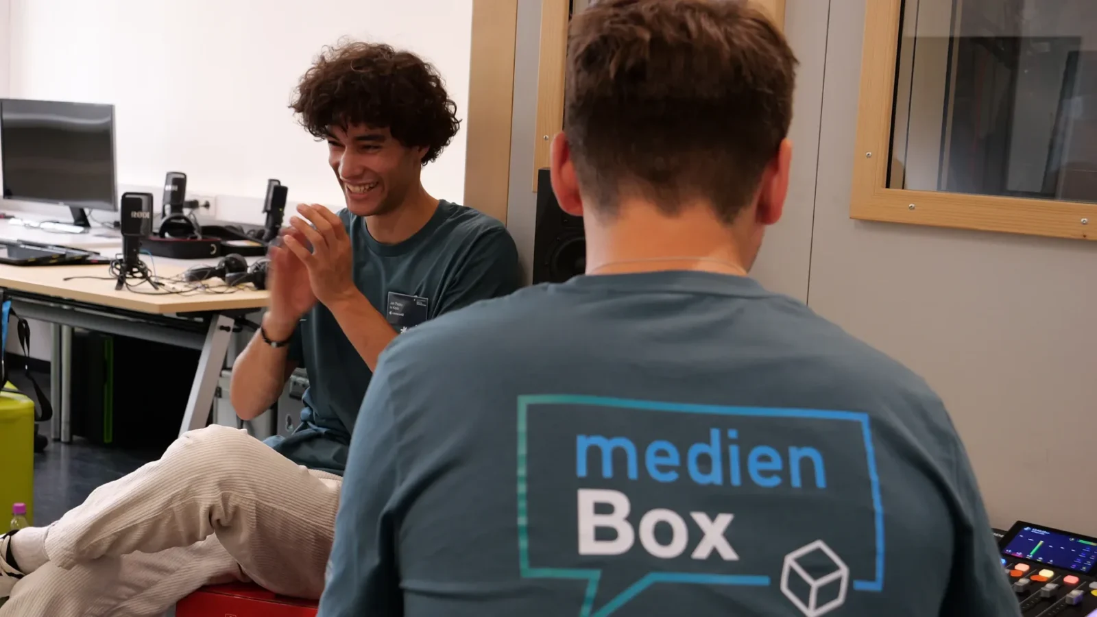 Lachender Mitarbeiter klatscht in die Hände zum Ton-Sync, davor ein Kollege am Mischpult