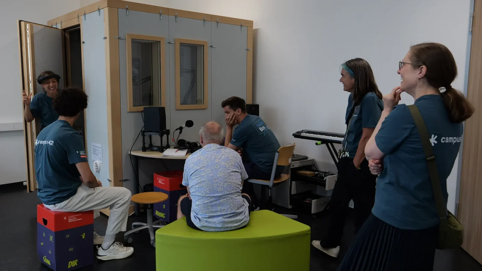 Gruppe im Podcast-Studio vor der Schallschutzkabine, Besucher auf Papphockern „Gönn dir Bildung"
