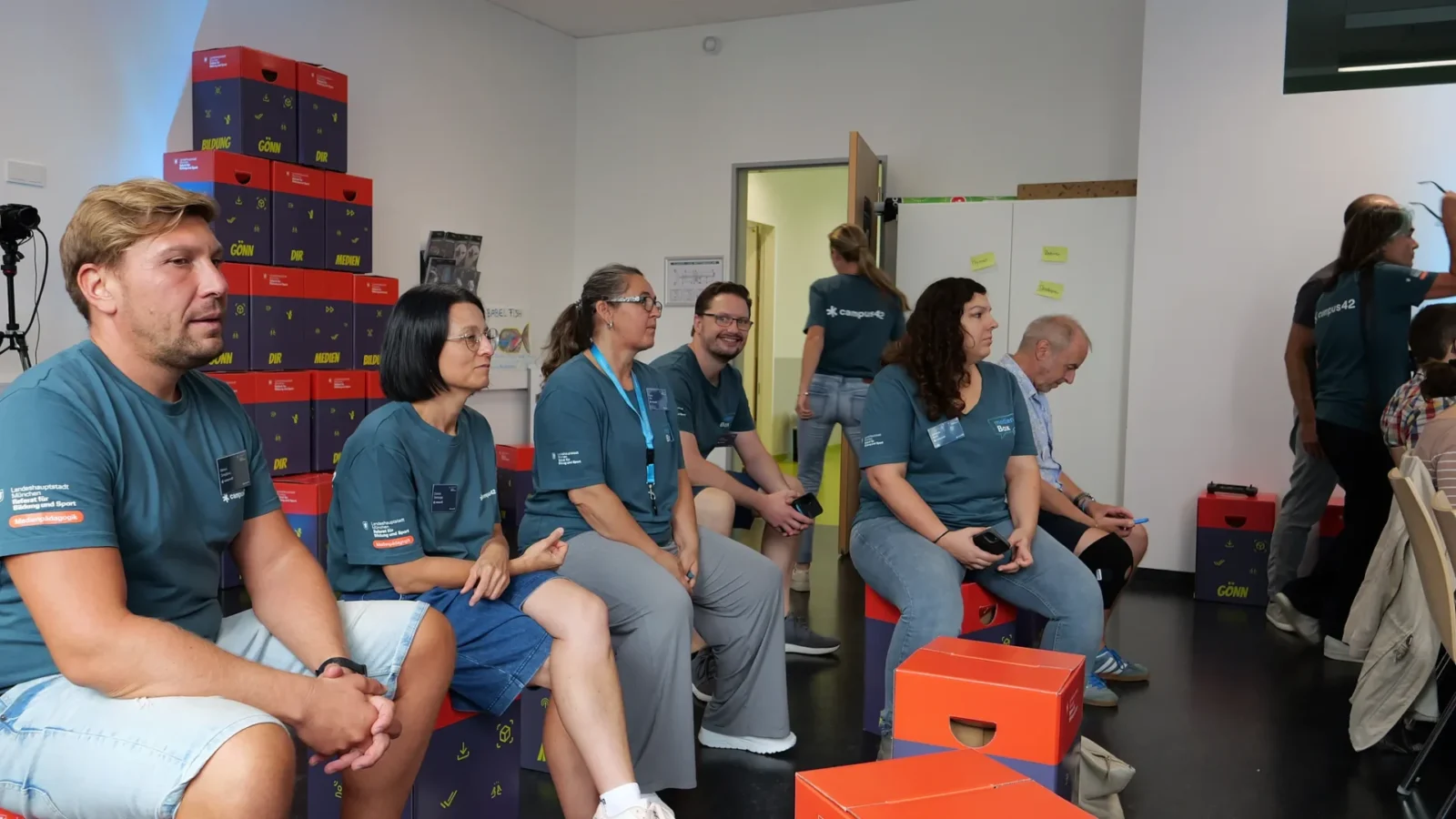 Teammitglieder sitzen auf gestapelten medienBox-Kisten mit dem Aufdruck „Gönn dir Medien Bildung"