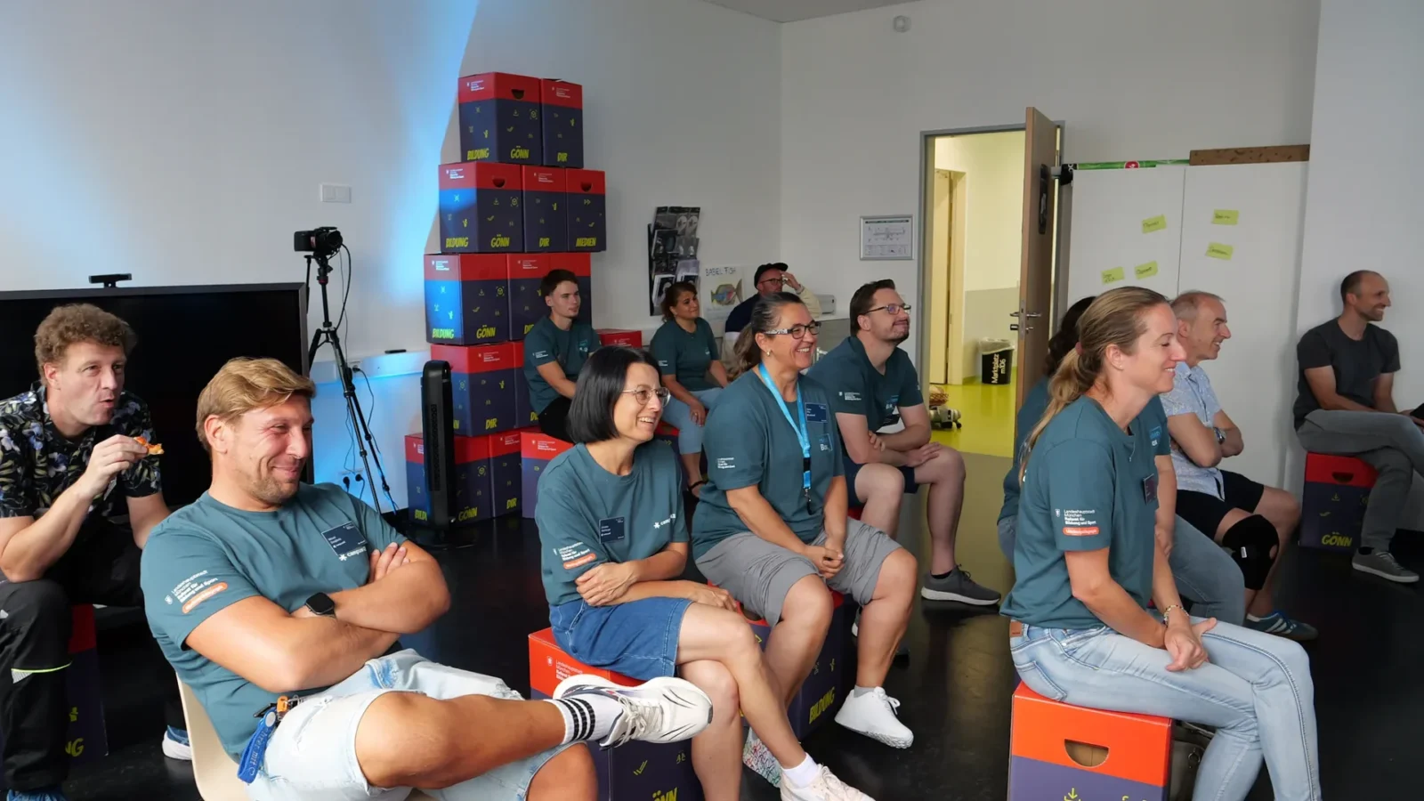 Lachendes Team auf medienBox-Kisten vor der Kistenpyramide „Bildung – Gönn dir – Medien"