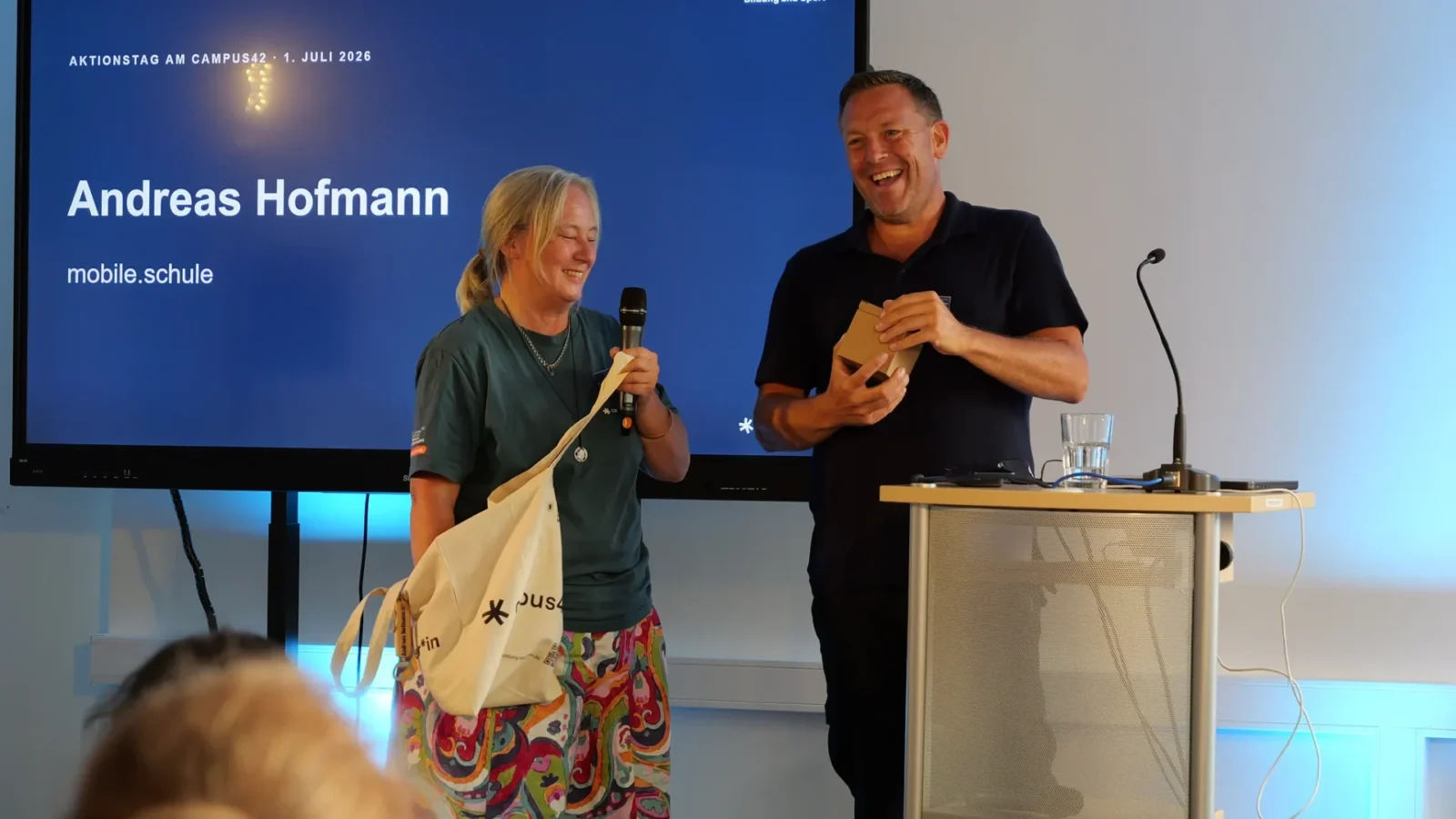 Lachende Moderatorin mit Mikrofon und campus42-Stofftasche neben dem Referenten