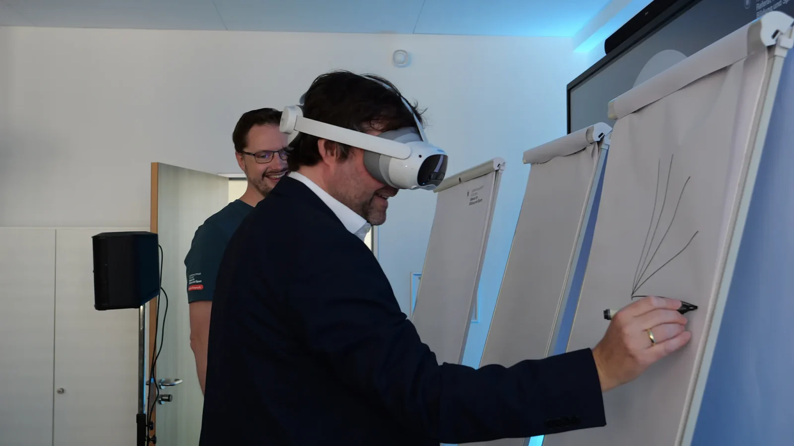 Gast malt mit VR-Brille und Stift auf einem Flipchart, ein Mitarbeiter schaut zu