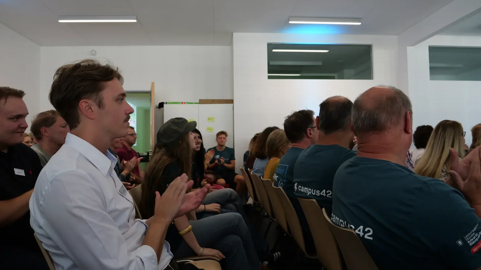 Applaudierendes Publikum auf Stuhlreihen, im Vordergrund Team-Mitglieder in campus42-Shirts