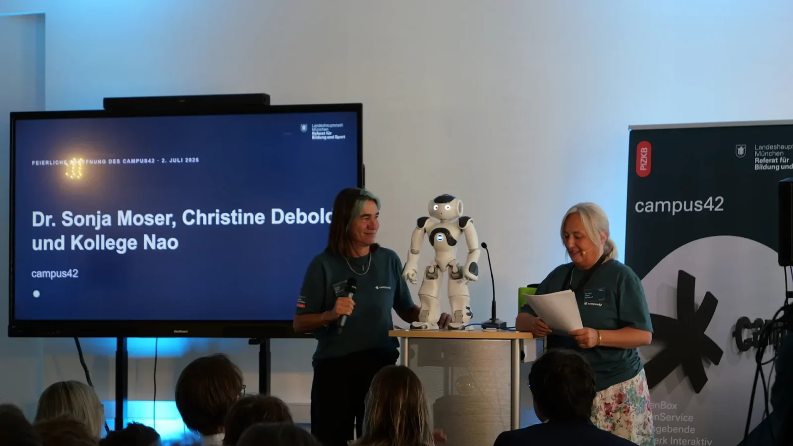 Zwei Moderatorinnen in campus42-Shirts präsentieren den humanoiden Roboter Nao