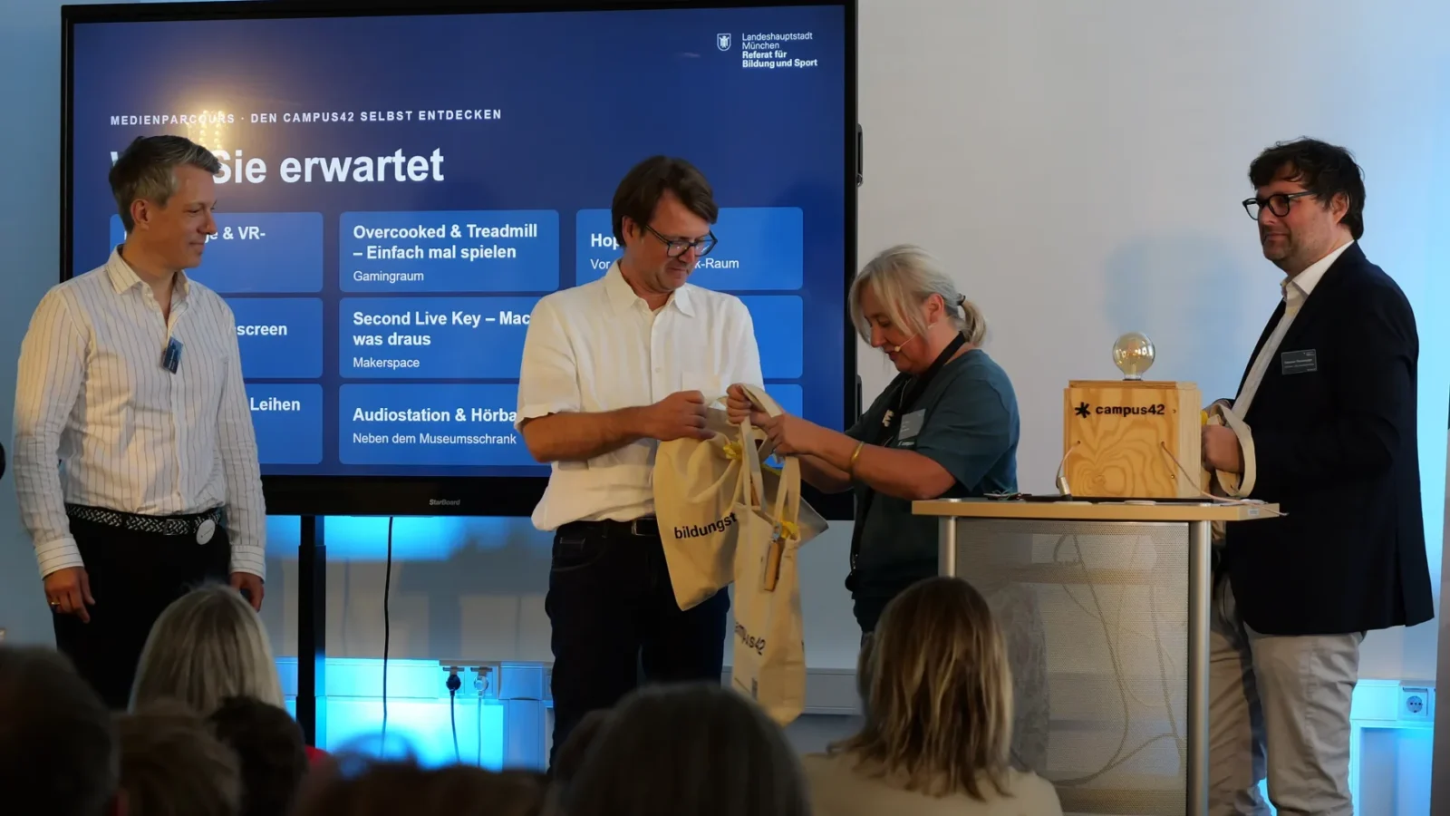 Übergabe einer Jutetasche vor dem Bildschirm mit der Programmübersicht
