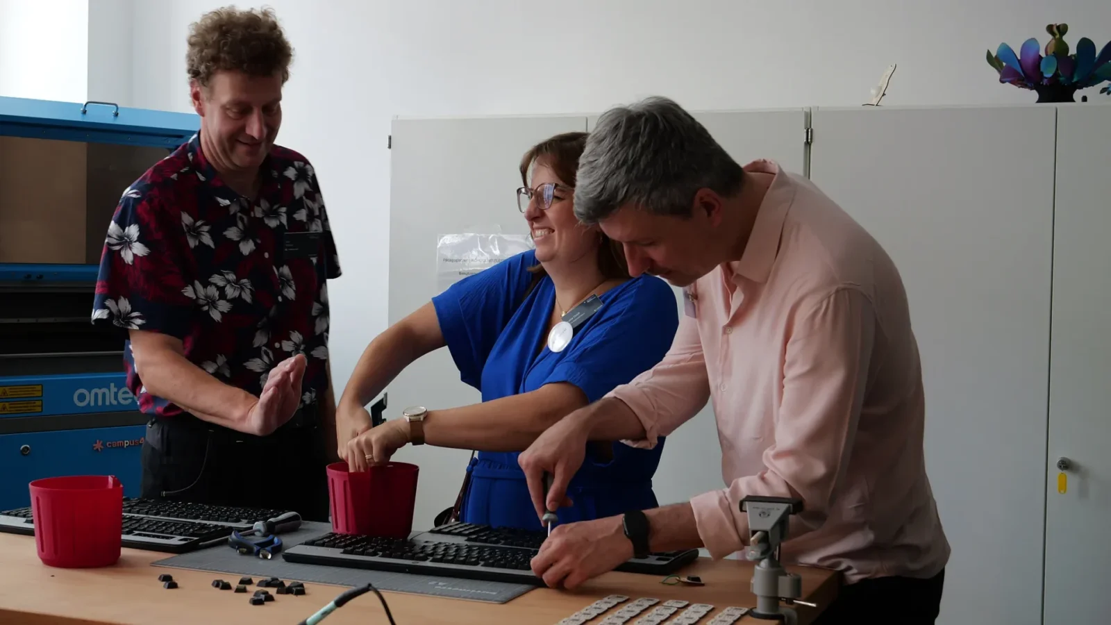 Drei Erwachsene lachen an der Werkbank im Makerspace, im Hintergrund der Lasercutter