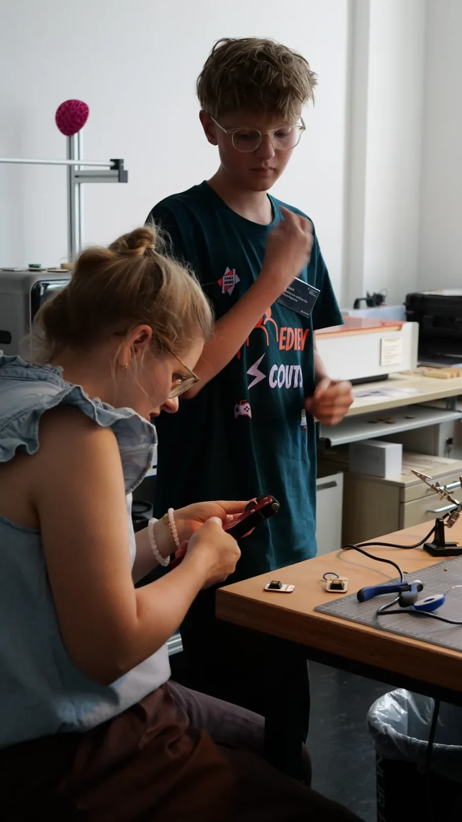 Zwei Personen bearbeiten Bauteile am Werktisch, im Hintergrund der Lasercutter