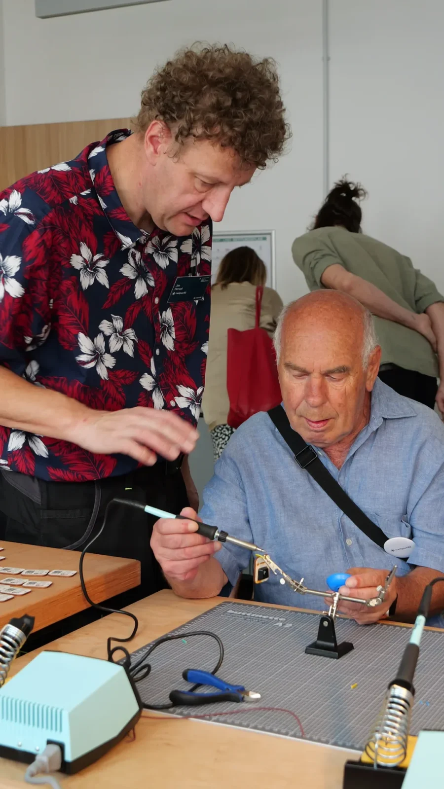 Ein Mitarbeiter zeigt einem älteren Besucher, wie ein Bauteil mit der Dritten Hand gelötet wird