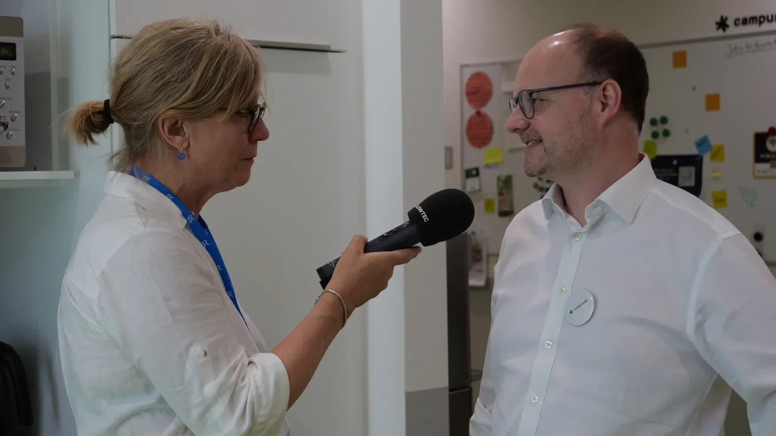 Radioreporterin mit BR-Mikrofon interviewt einen Mann mit campus42-Button vor der Pinnwand
