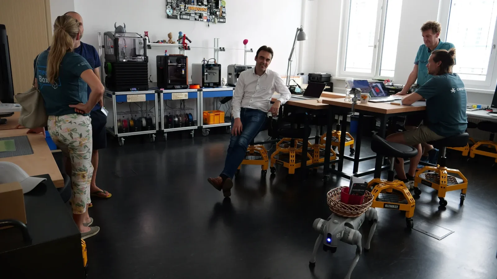 Makerspace mit 3D-Druckern, Filamentrollen und Roboterhund; Team im Gespräch an den Werkbänken