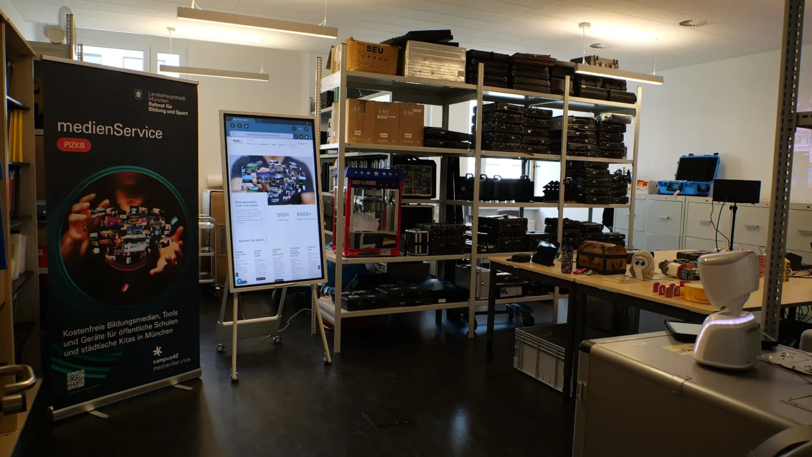 Materiallager des medienService mit Rollup, Touchdisplay und Regalen voller Technikkoffer und Geräte
