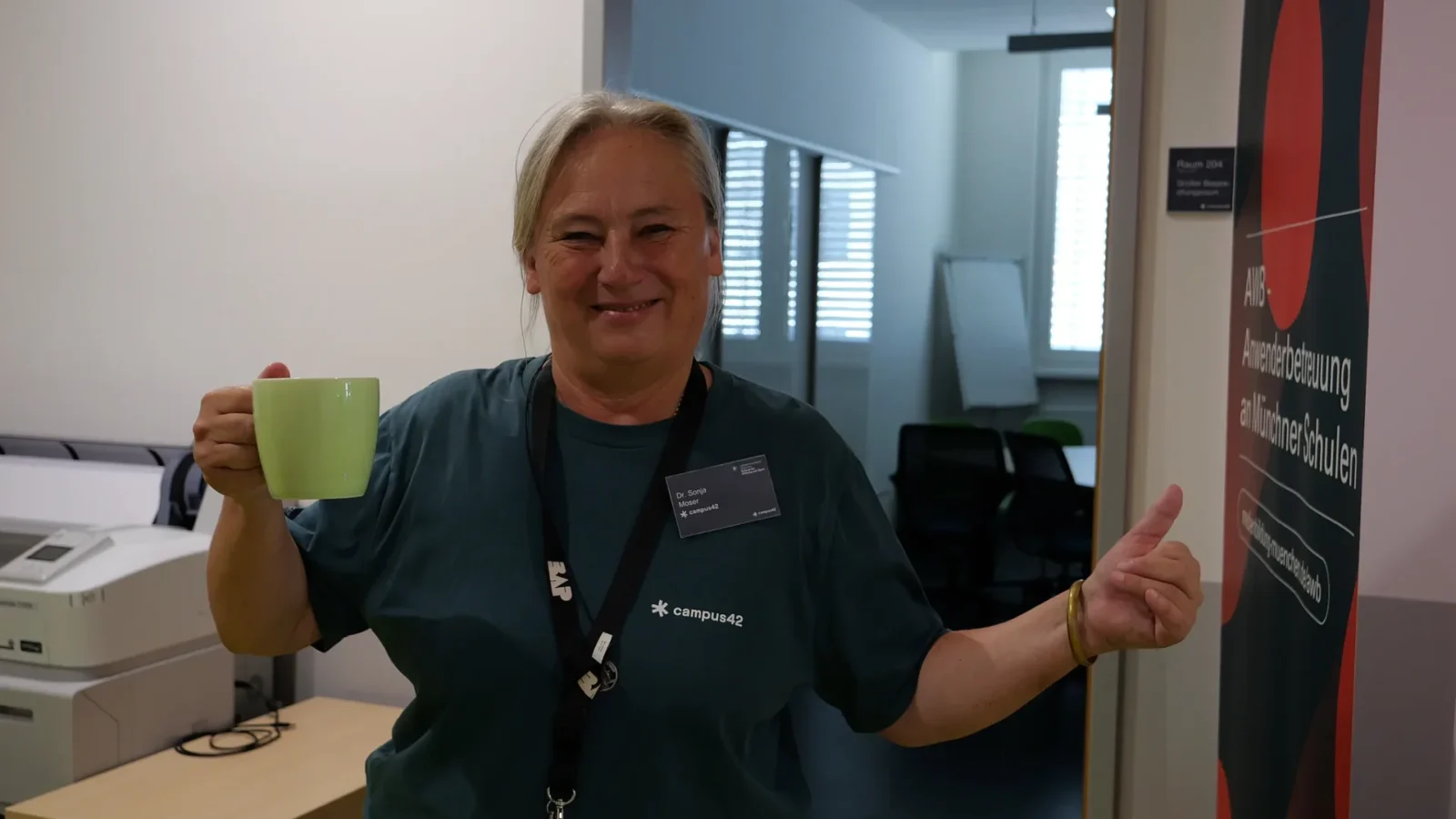 Mitarbeiterin mit campus42-Shirt hebt eine Tasse und zeigt Daumen hoch vor dem Rollup