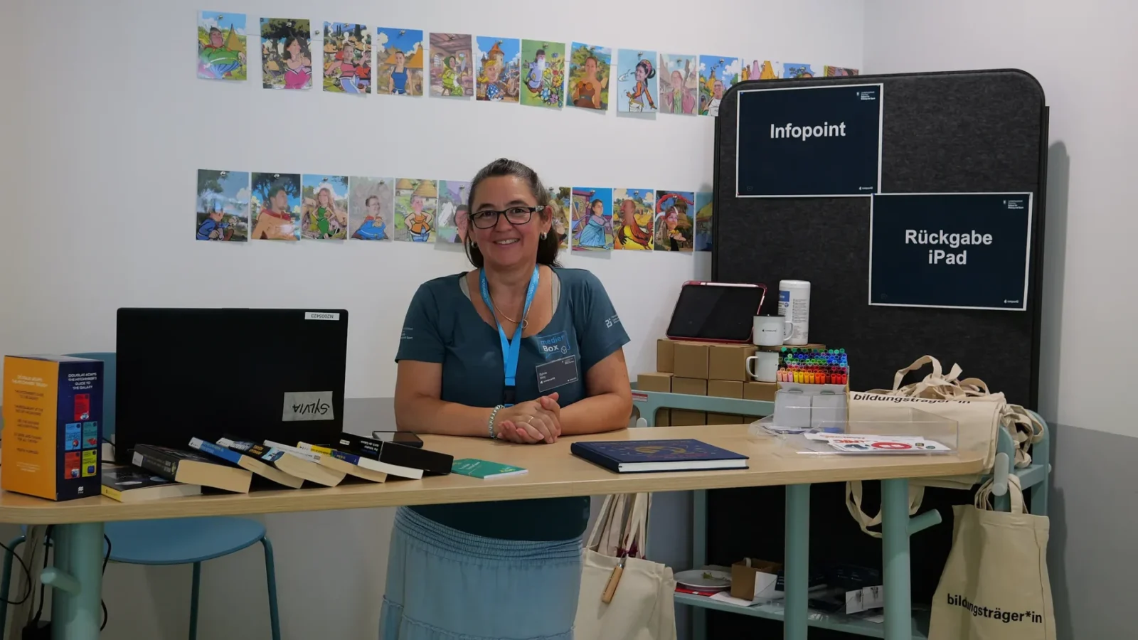 Infopoint mit iPad-Rückgabe, Bücherauslage und Stiften, dahinter eine Wand mit Comic-Zeichnungen