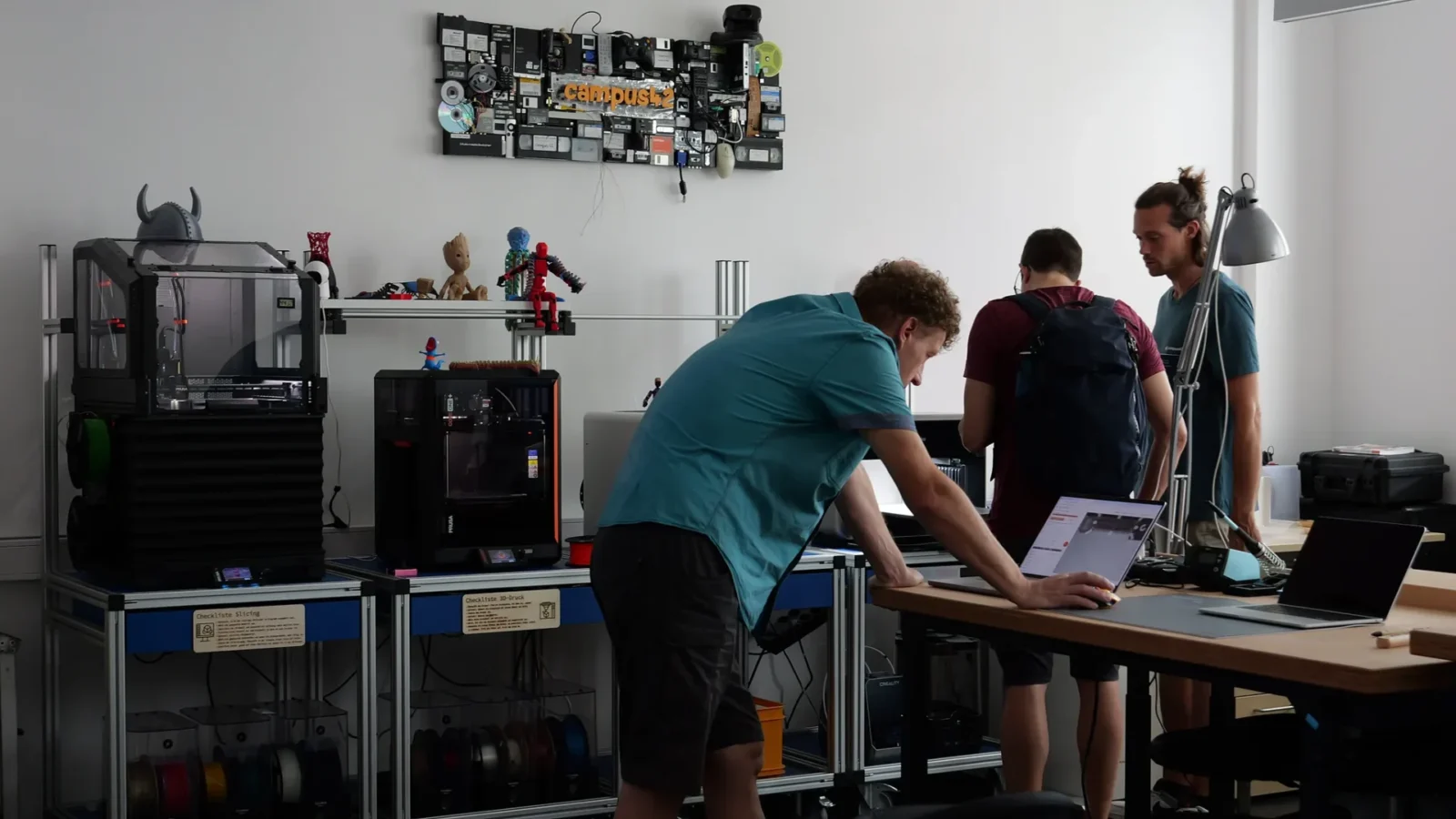 Makerspace mit Prusa-3D-Druckern, Filamentrollen und Druckobjekten; Mitarbeitende am Laptop