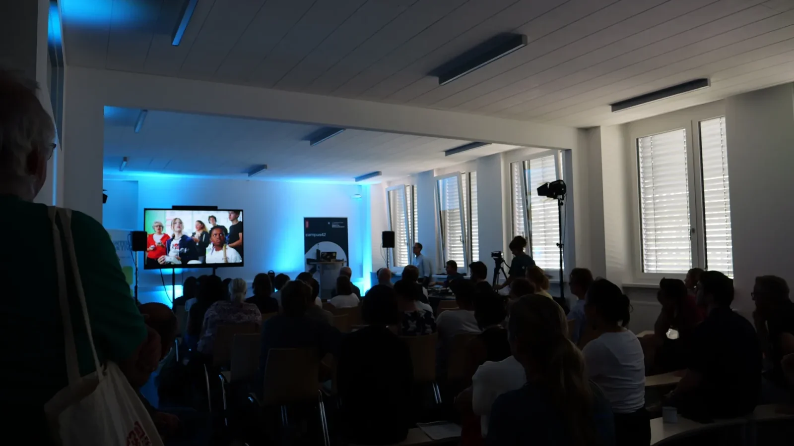 Publikum im abgedunkelten Veranstaltungsraum verfolgt eine Filmpräsentation auf großem Bildschirm