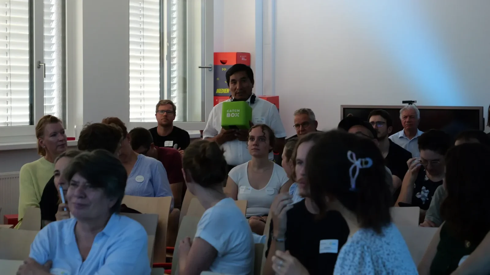 Wortmeldung im Publikum mit dem grünen Catchbox-Wurfmikrofon, dahinter Kisten mit Aufschrift „Medien"