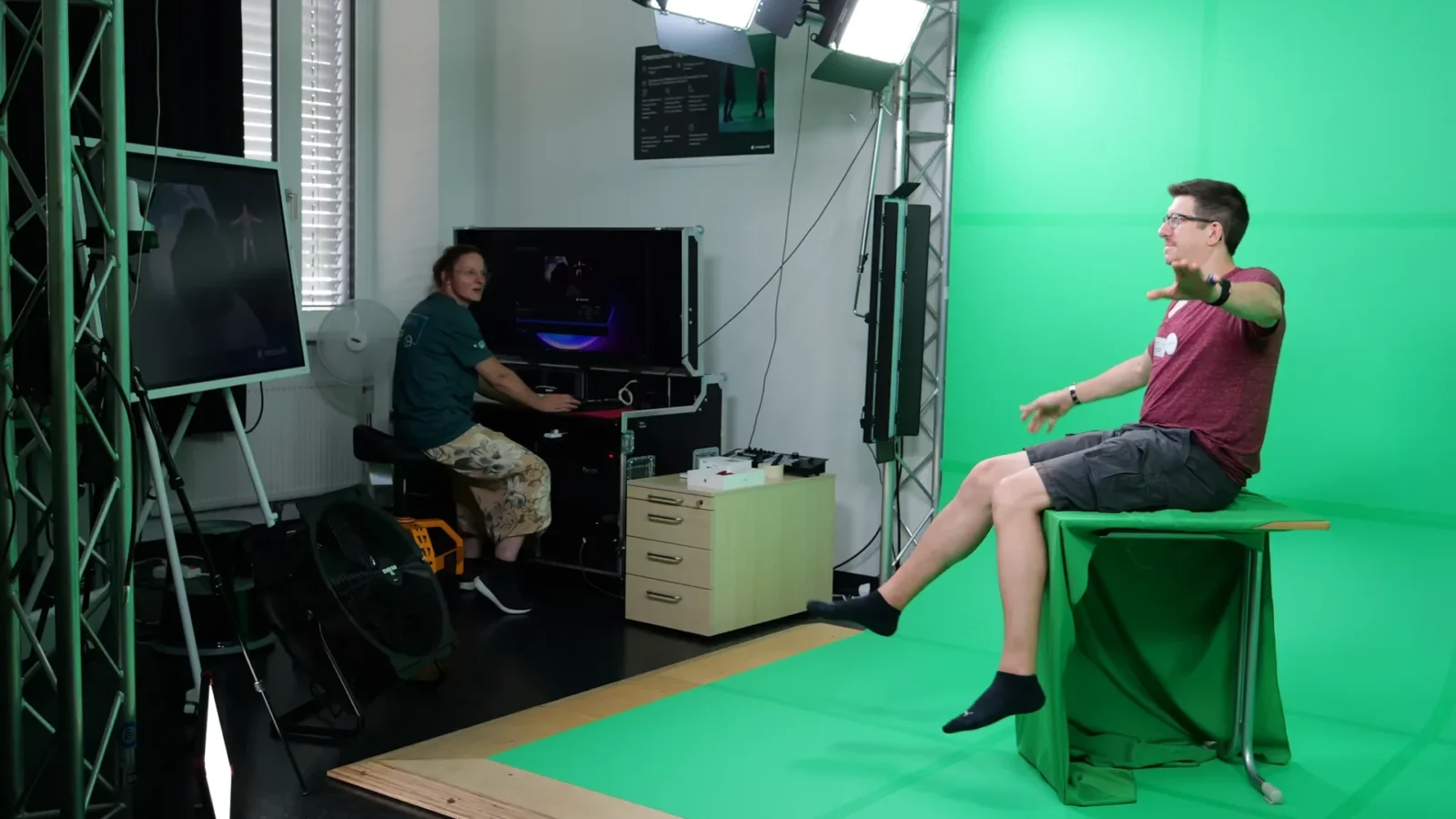 Greenscreen-Filmstudio: Person sitzt gestikulierend vor der Greenscreen-Wand, Technikerin steuert den Schnittplatz